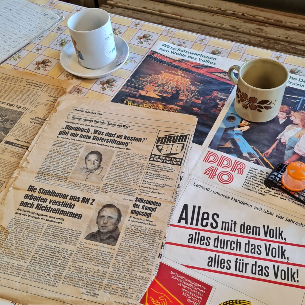 Ein Tisch auf dem Seiten aus Zeitungen und Zeitschriften ausgebreitet sind. Die oberste Zeitungsseite enthält Artikel mit den Überschriften: "Handbuch 'Was darf es kosten?' gibt mir gute Unterstützung", "Die Stahlbauer aus RH 2 arbeiten verstärkt nach Richtzeitnormen", "Stillständen der Kampf angesagt".Daneben eine Zeitschriftenseite, auf der zu lesen ist: "Leitmotiv unseres Handelns seit über vier Jahrzehnten. Alles mit dem Volk, alles durch das Volk, alles für das Volk!"