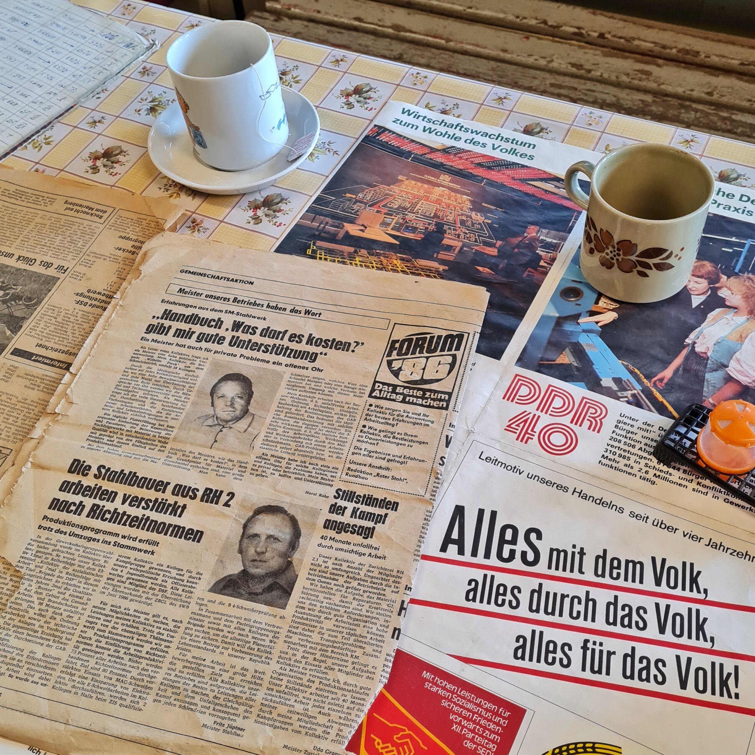 Ein Tisch auf dem Seiten aus Zeitungen und Zeitschriften ausgebreitet sind. Die oberste Zeitungsseite enthält Artikel mit den Überschriften: "Handbuch 'Was darf es kosten?' gibt mir gute Unterstützung", "Die Stahlbauer aus RH 2 arbeiten verstärkt nach Richtzeitnormen", "Stillständen der Kampf angesagt".Daneben eine Zeitschriftenseite, auf der zu lesen ist: "Leitmotiv unseres Handelns seit über vier Jahrzehnten. Alles mit dem Volk, alles durch das Volk, alles für das Volk!"