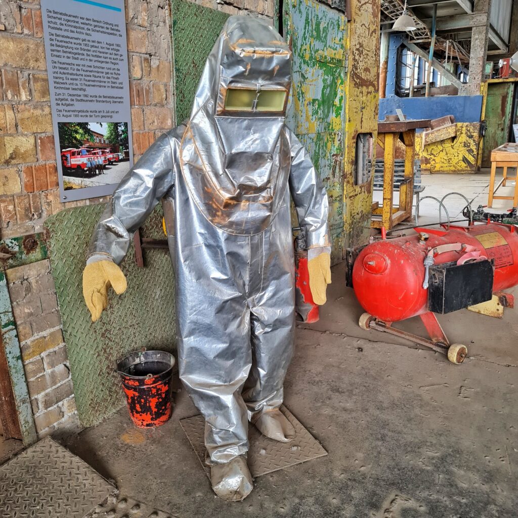 Eine mannsgroße Figur, die einen weiß beschichteten Schutzanzug trägt. Dieser bedeckt den ganzen Körper und hat vor dem Gesischt eine eingearbeitete durchsichtige Schutzbrille. An den Händen hat die Figur dicke Handschuhe.Neben ihr ein Eimer. An der Wamd neben ihr ist ein Teil eines Erklärungstextes mit einem Foto darunter zu sehen. Das Foto zeigt Feuerwehrfahrzeuge.
