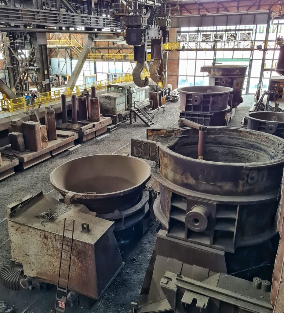 Ein anderer Blickwinkel. Im Vordergrund die riesigen Gefäße. Im Hintergrund die Lok, die beiden Haken, die von der Decke hängen und viele von den hohen , viereckigen Metallgefäßen.