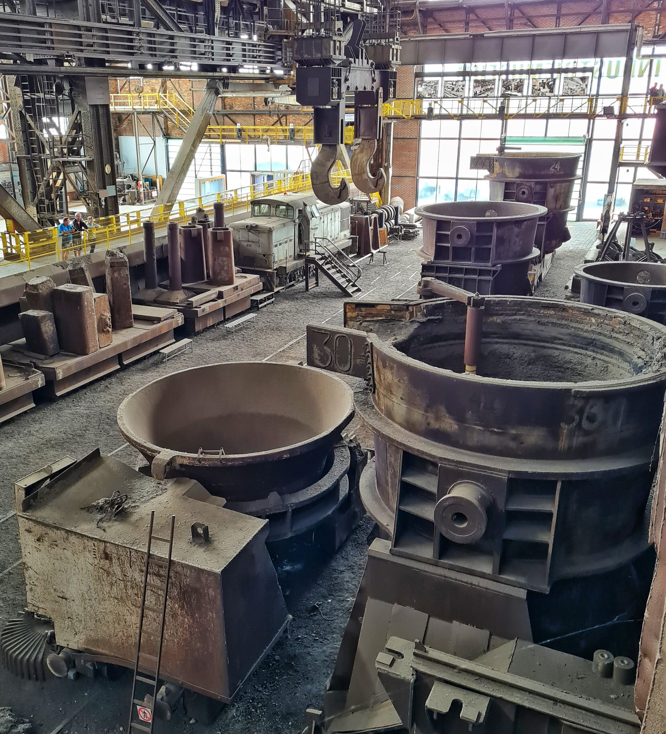 Ein anderer Blickwinkel. Im Vordergrund die riesigen Gefäße. Im Hintergrund die Lok, die beiden Haken, die von der Decke hängen und viele von den hohen , viereckigen Metallgefäßen.
