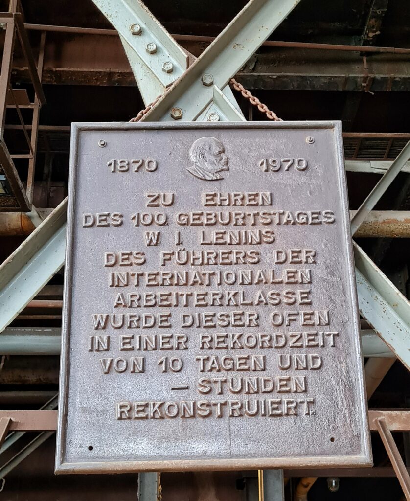 Eine Metalltafel, die an querstehenden Metallstreben angebracht ist. Auf ihr steht: 1970 -1980. Zu Ehren des 100. Geburtstages W. I. Lenin, des Führers der deutschen Arbeiterklasse, wurde dieser Ofen in einer Rekordzeit von 10 Tagen und - Stunden rekonstruiert.