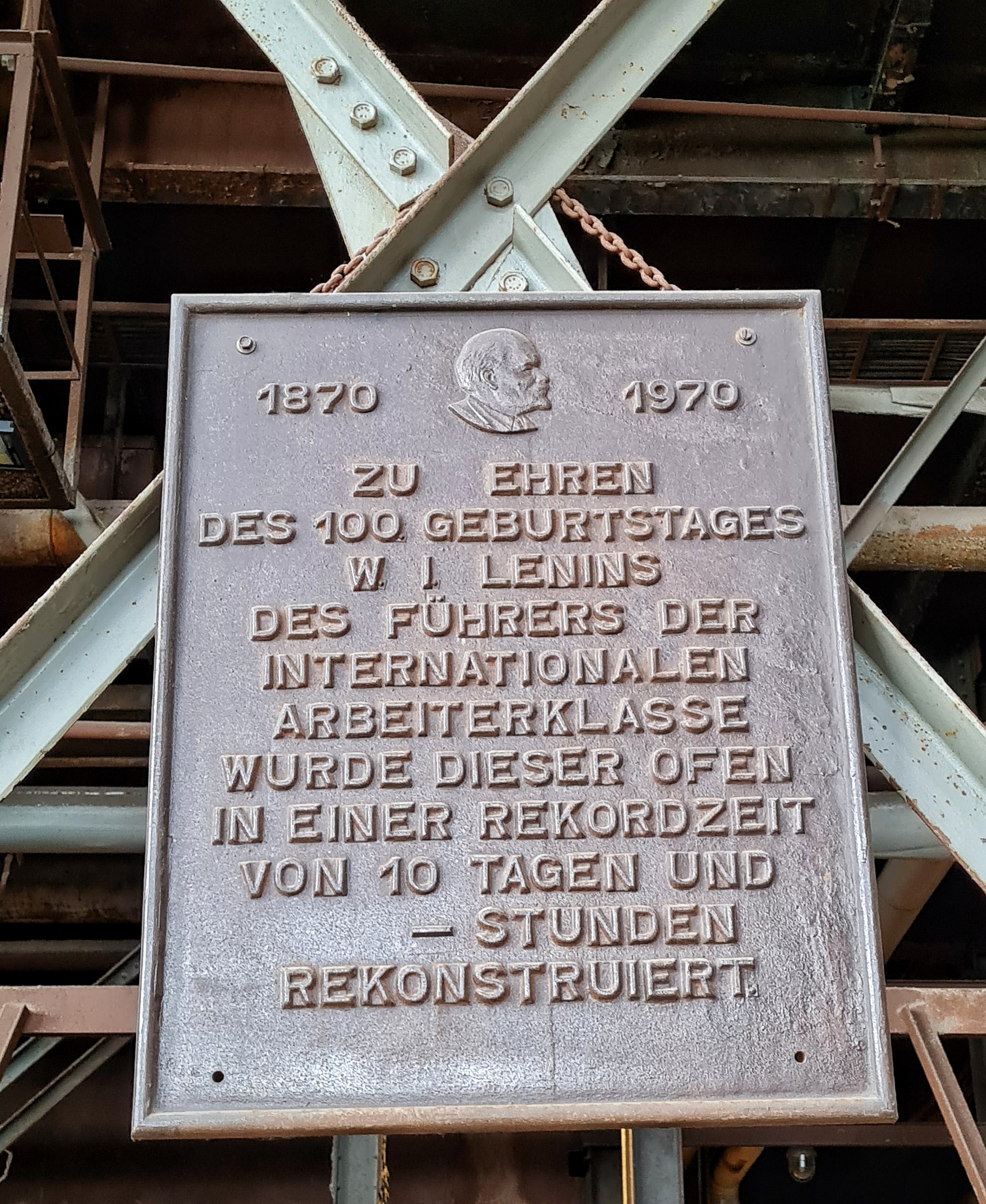 Eine Metalltafel, die an querstehenden Metallstreben angebracht ist. Auf ihr steht: 1970 -1980. Zu Ehren des 100. Geburtstages W. I. Lenin, des Führers der deutschen Arbeiterklasse, wurde dieser Ofen in einer Rekordzeit von 10 Tagen und - Stunden rekonstruiert.