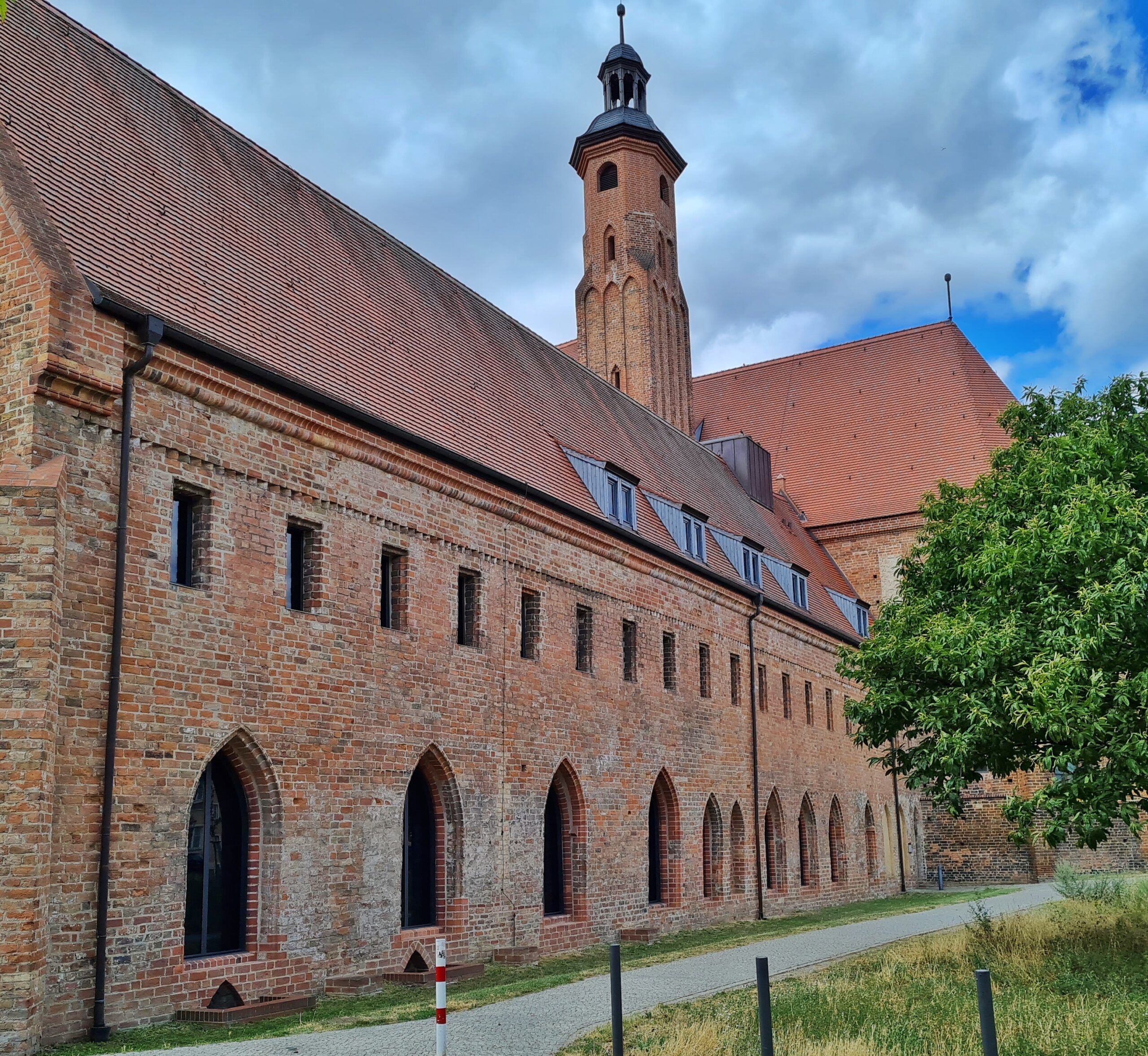 Im Vordergrund ein längliches Backsteingebäude. Unten mehrere halbrunde Öffnungen nebeneinander. Oben sehr kleine schmale Öffnungen nebeneinander. Am hinteren Teil des Gebäudes schließt sich ein quergestelltes Gebäude