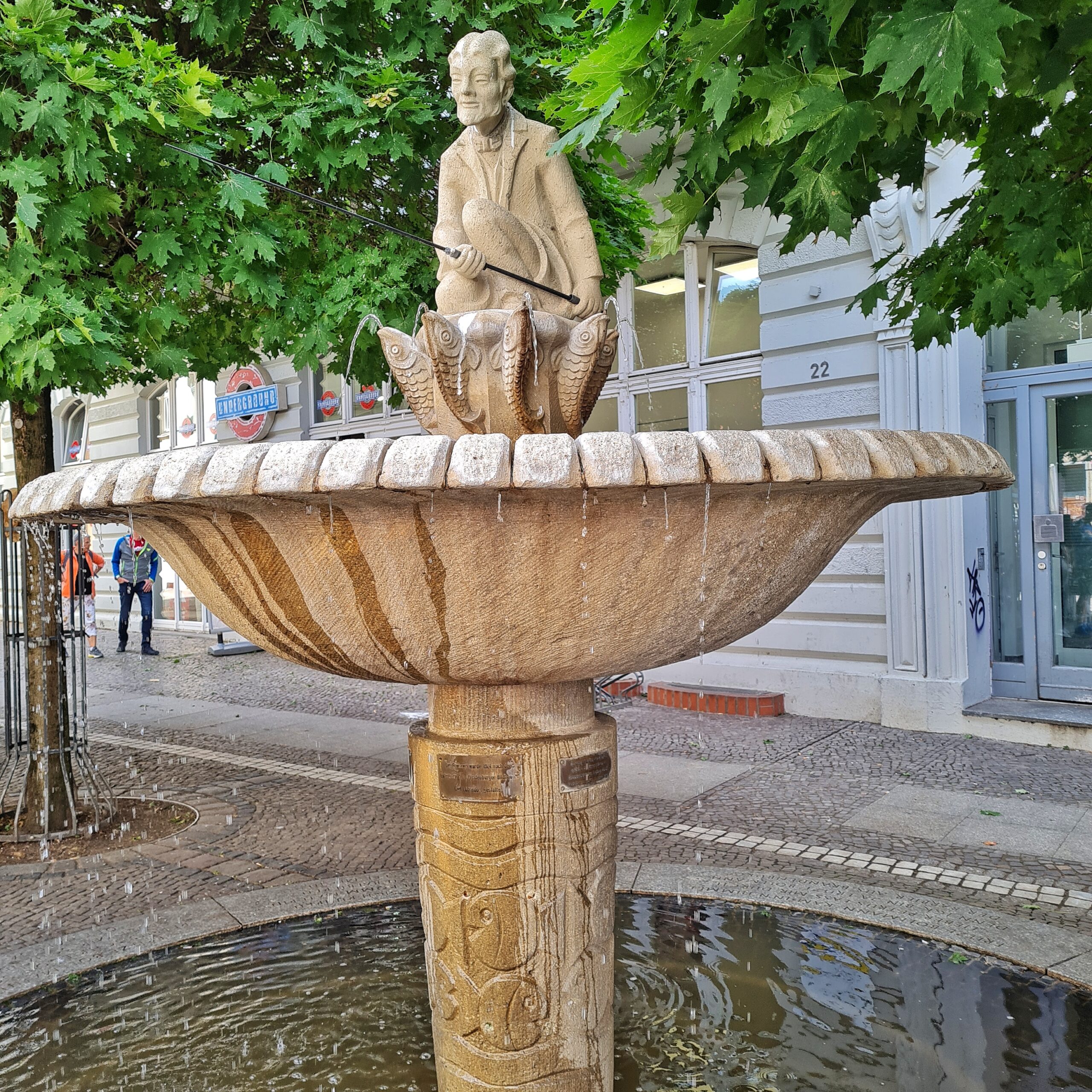 Ein heller Brunnen in einer Straße mit Geschäften und laufenden Menschen. Oben die Figur eines älteren hockenden Mannes mit einer Angel in der Hand. Er sitzt auf einem kleinen Podest, an dem außen senkrecht Fische nach oben ragen, aus deren Mäulern Wasser spritzt. Das Wasser spritzt in eine große Schale, aus der wiederum Wasser in ein noch größeres Wasserbecken am Boden tropft.