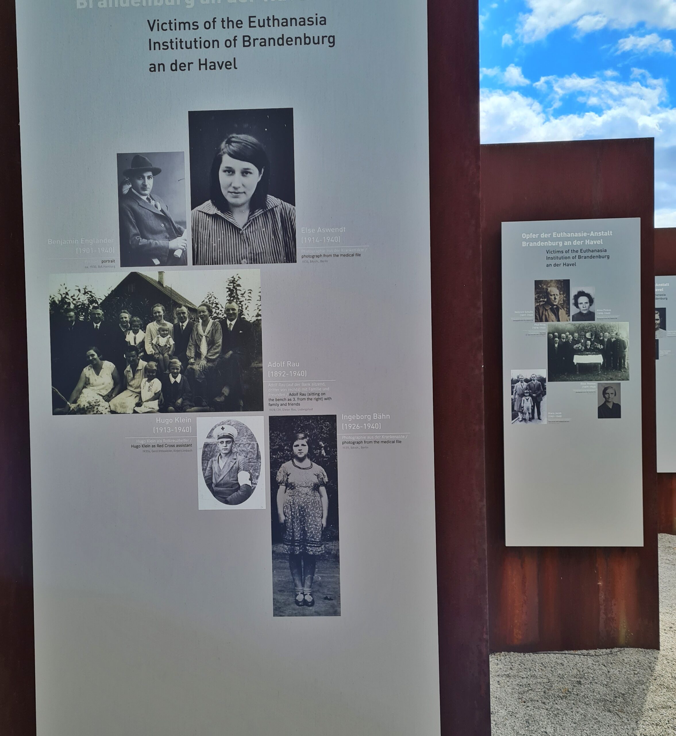 Mit einigem Abstand stehen hintereinander große Metallplatten. Darauf alte Schwarz-Weiß-Fotos und daneben kurzer Text. Auf der vorderen Platte steht oben: Victims of the Euthanasia Institution of Brandenburg an der Havel.