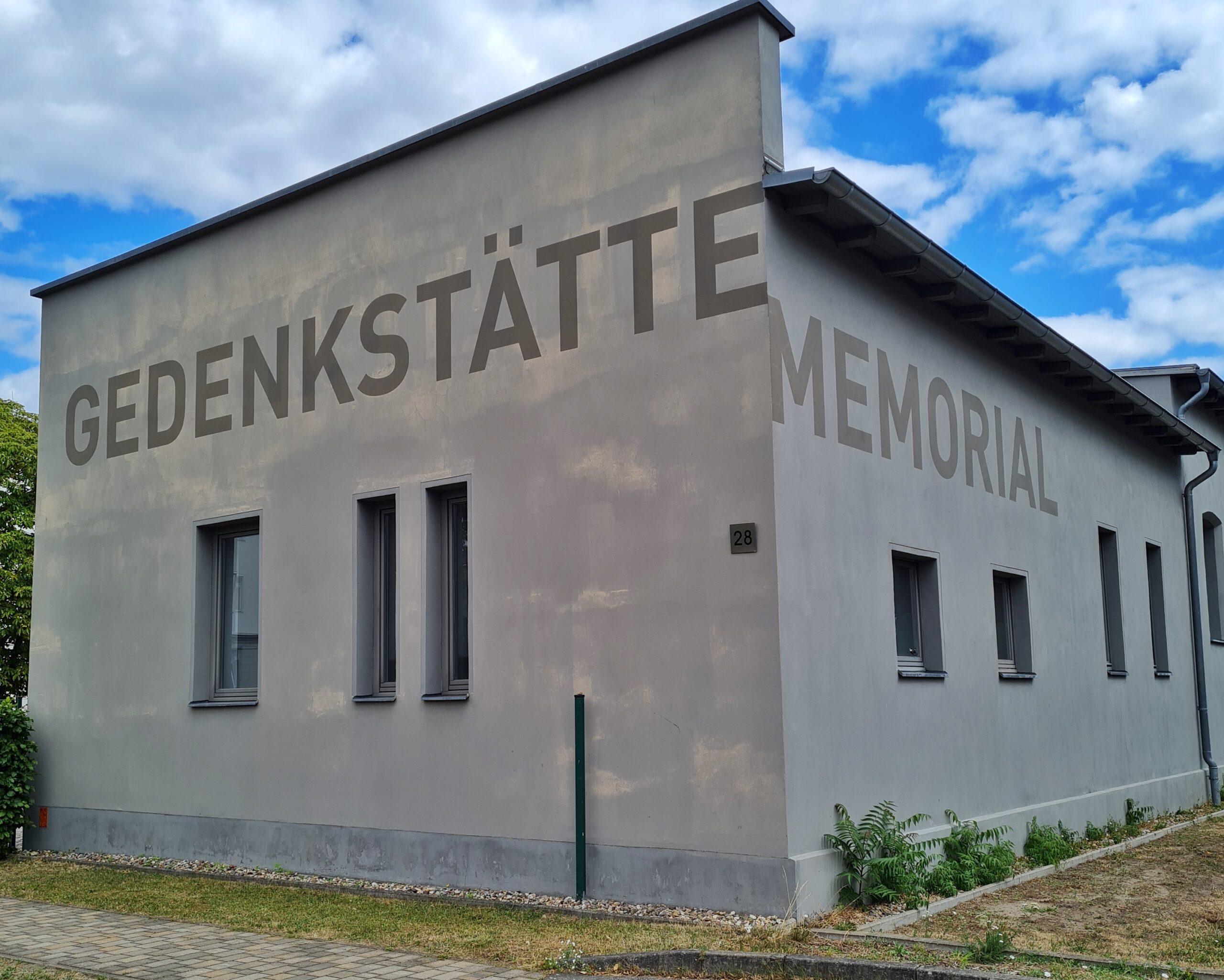 Ein hellgraues niedriges Gebäude mit einigen schmalen Fenstern. Auf der einen Seite steht in großen Buchstaben: Gedenkstätte, auf der anderen: Memorial.
