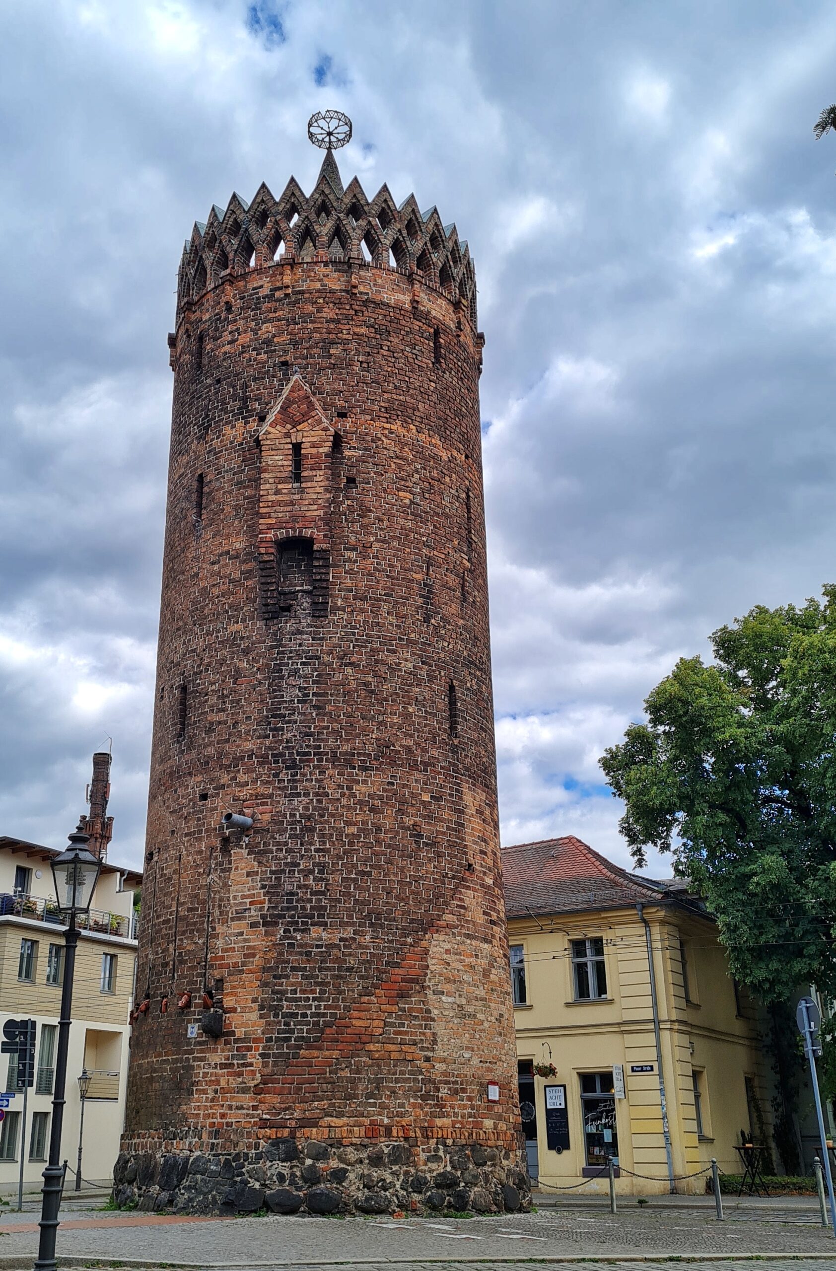 Ein hoher runder Backsteinturm vor einigen hellen Gebäuden mit Wohnungen und Läden. Er hat nur wenige schmale Öffnungen. Oben eine Umrandung aus zwei gezackten Linien. Darin ein radförmiger Gegenstand auf einer Spitze.