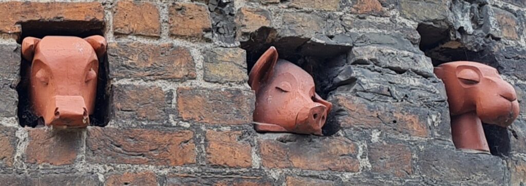 Herangezoomt eine Nische in einer Backsteinmauer. Darin liegt der Kopf eines schlafenden Schweines aus Ton oder ähnlichem Material.