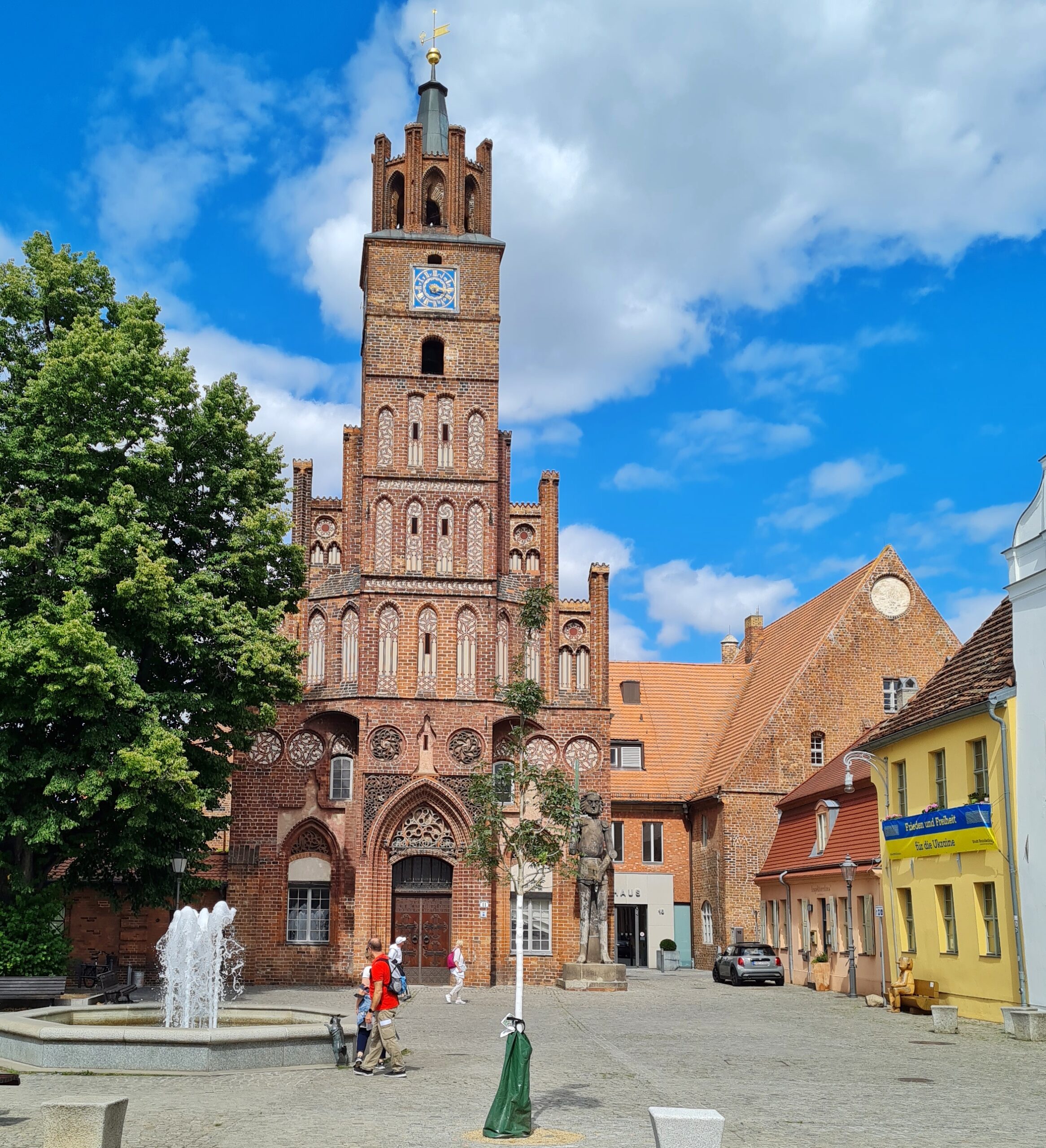 Ein Platz mt einem Springbrunnen und einem großen Baum. Dahinter ein kirchenähnliches Gebäude aus Backstein. Es ist unten etwas breiter und verengt sich dann in einen Turm mit mehreren Etagen. Rechts davon steht eine große männliche Statue , die ein Schwert hochhält. Rechts weitere niedrigere Backsteingebäude.