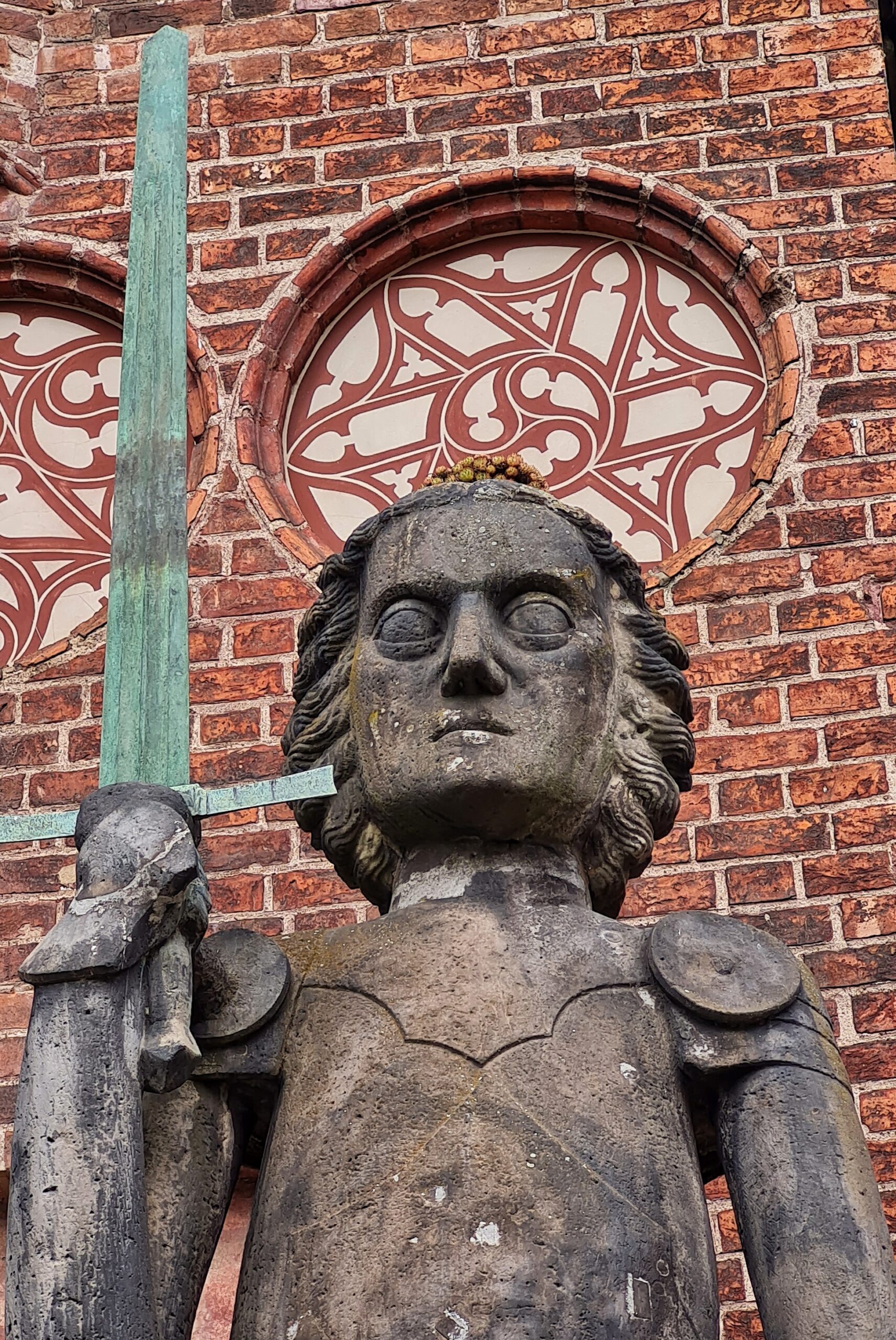 Der Oberkörper, die Hand mit dem Schwert und der Kopf der Statue herangezoomt. Auf dem Kopf wächst eine niedrige Pflanze.