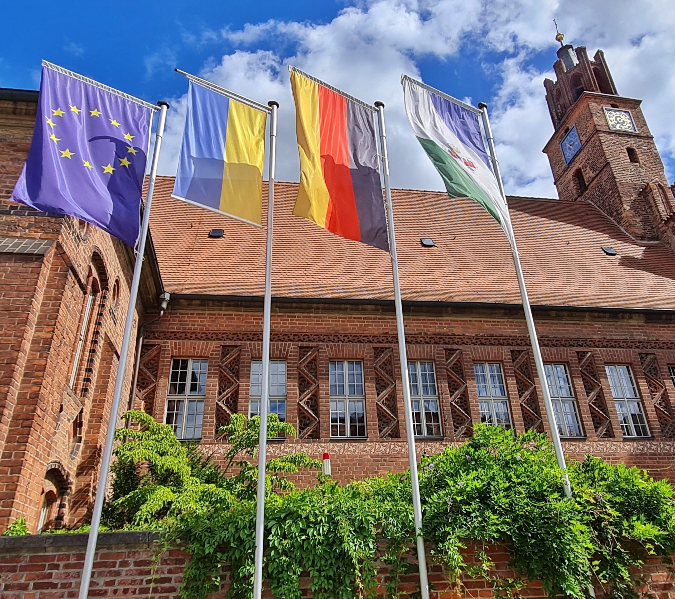 Das Gebäude in der Seitenansicht. Die Mauer unten ist grün bewachsen. Im Obergeschoß mehrere Sprossenfenster, dazwischen zackenförmige Verzierungen. Vor dem Gebäude wehen an hohen Stangen die Europaflagge, die Flagge der Ukraine, die Deutschlandflagge und die Flagge der Stadt Brandenburg an der Havel.