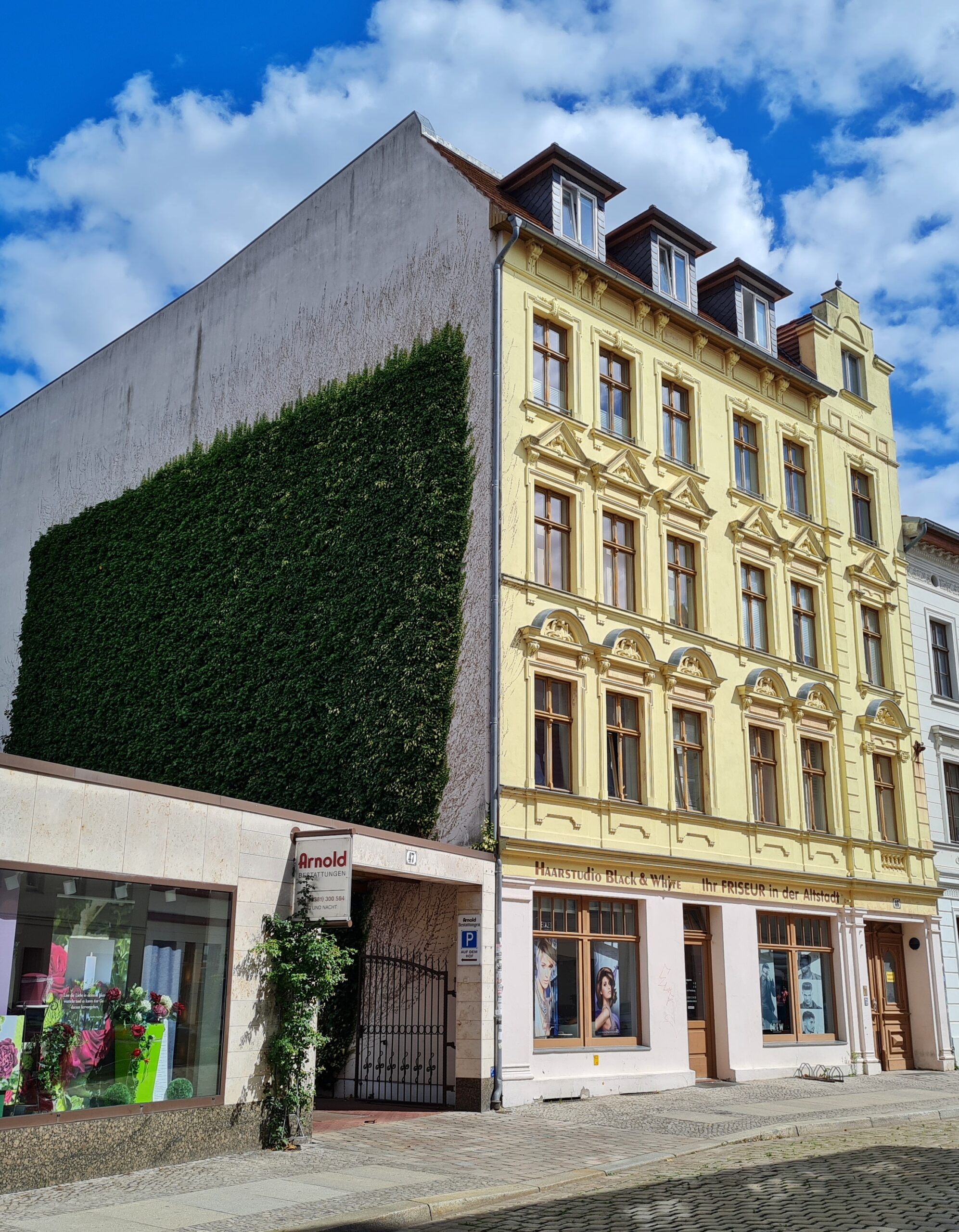 Ein hohen helles Gebäude mit Sprossenfenstern, Verzierungen und Dachgauben. Die linke Wand ist bis zu einem flachen Gebäude, in dem ein Bestattungsinstitut ist, begrünt.
