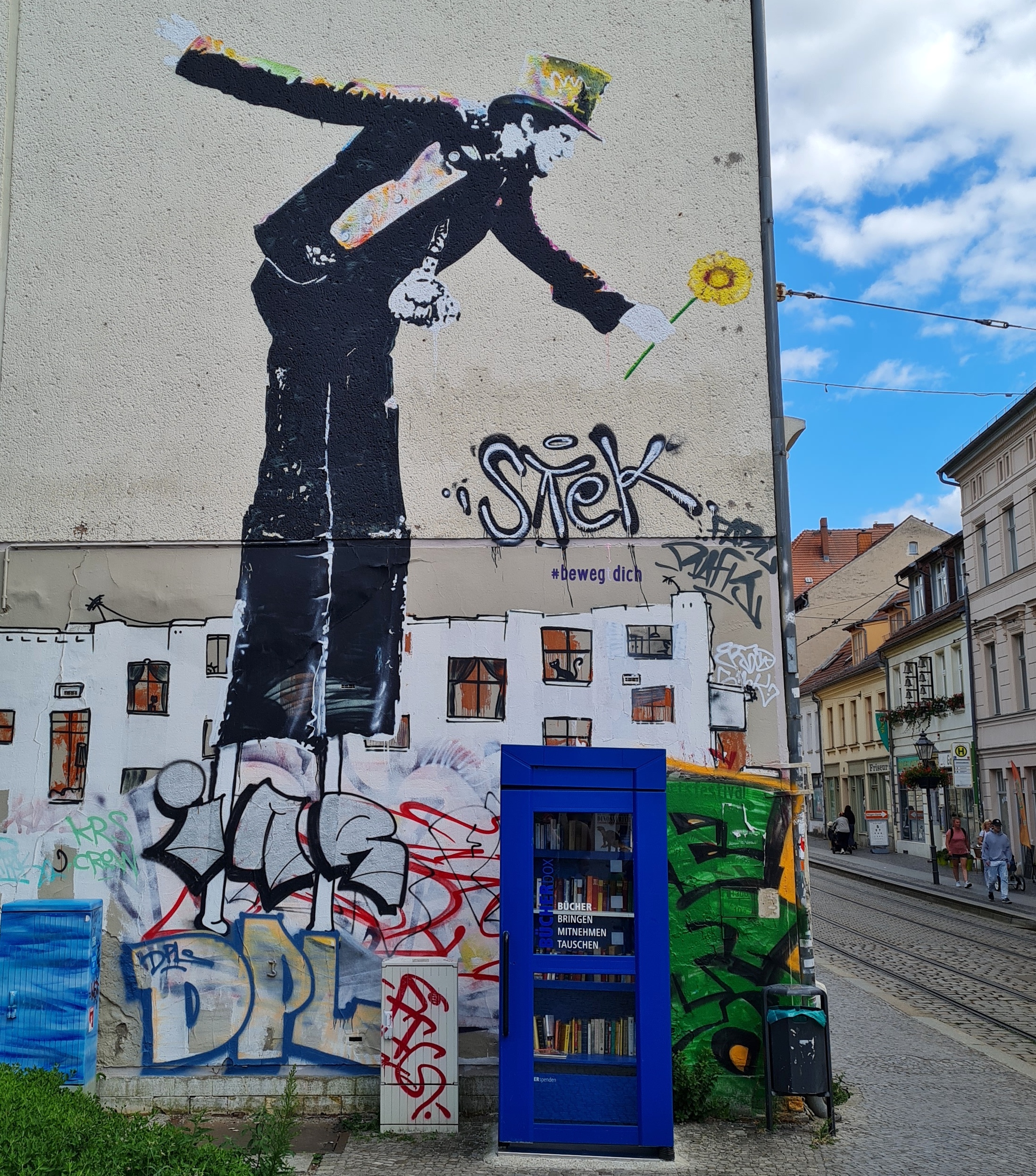 Eine Hauswand. rechts eine Straße mit Straßenbahngleisen und Geschäften. Oben auf der Hauswand die große Zeichnung eines Mannes im schwarzen Anzug und mit einem gelblichen Zylinder. Eine Hand hält er nach hinten ausgestreckt. Die andere Hand, in der er eine gelbe Blume hält, kst etwas nach unten gestreckt. Darunter die Zeichnung eines weißen Hauses mit vielen Fenstern, durch die man ins Innere schauen kann. Die Zeichnung wird jedoch teilweise durch große Graffiti-Schriftzüge verdeckt. Vor der Hauswand steht eine blaue Telefonzelle mit Büchern darin und der Aufschrift: Bücher bringen mitnehmen tauschen.