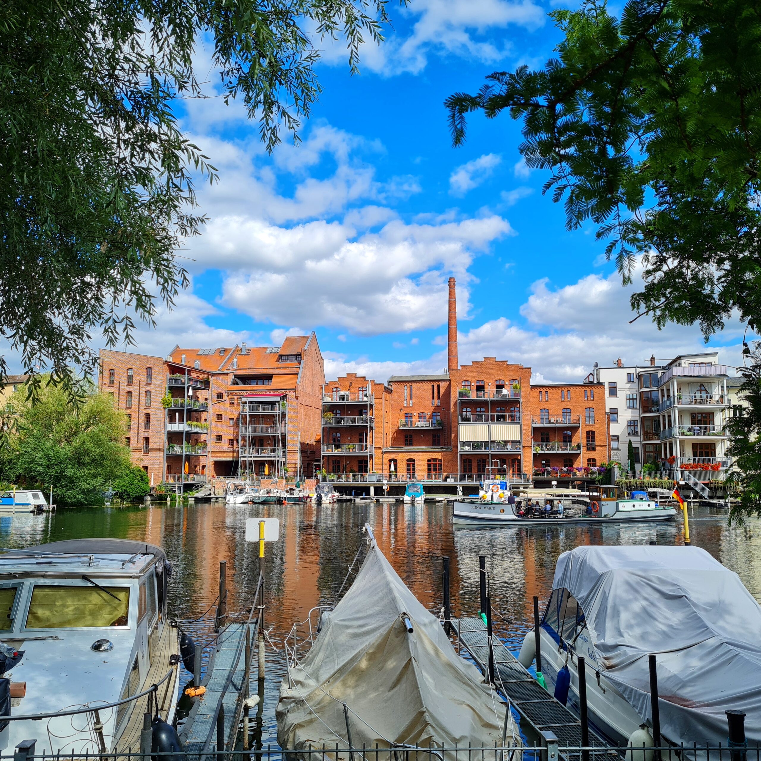 Blick über eine Wasserfläche auf moderne Wohngebäude mit Balkonen am anderen Ufer. In der Mitte fährt ein Ausflugsschiff. Vor dem Ufer, von dem aus fotografiert wurde, liegen drei größere Boote, teilweise mit weißen Planen abgedeckt. Links und rechts ragen Zweige mit grünen Blättern ins Bild.