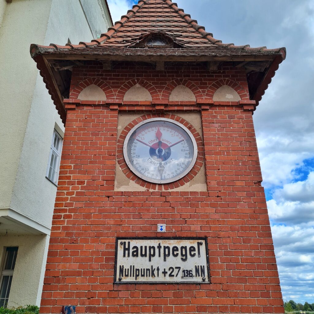 Ein roter Backsteinturm mit Dach. Oben ist hinter Glas eine runde Anzeige mit mehreren Zahlen und Zeigern. Darunter ein weißes Schild mit schwarzer Schrift: Hauptpegel. Nullpunkt + 27 136 NN