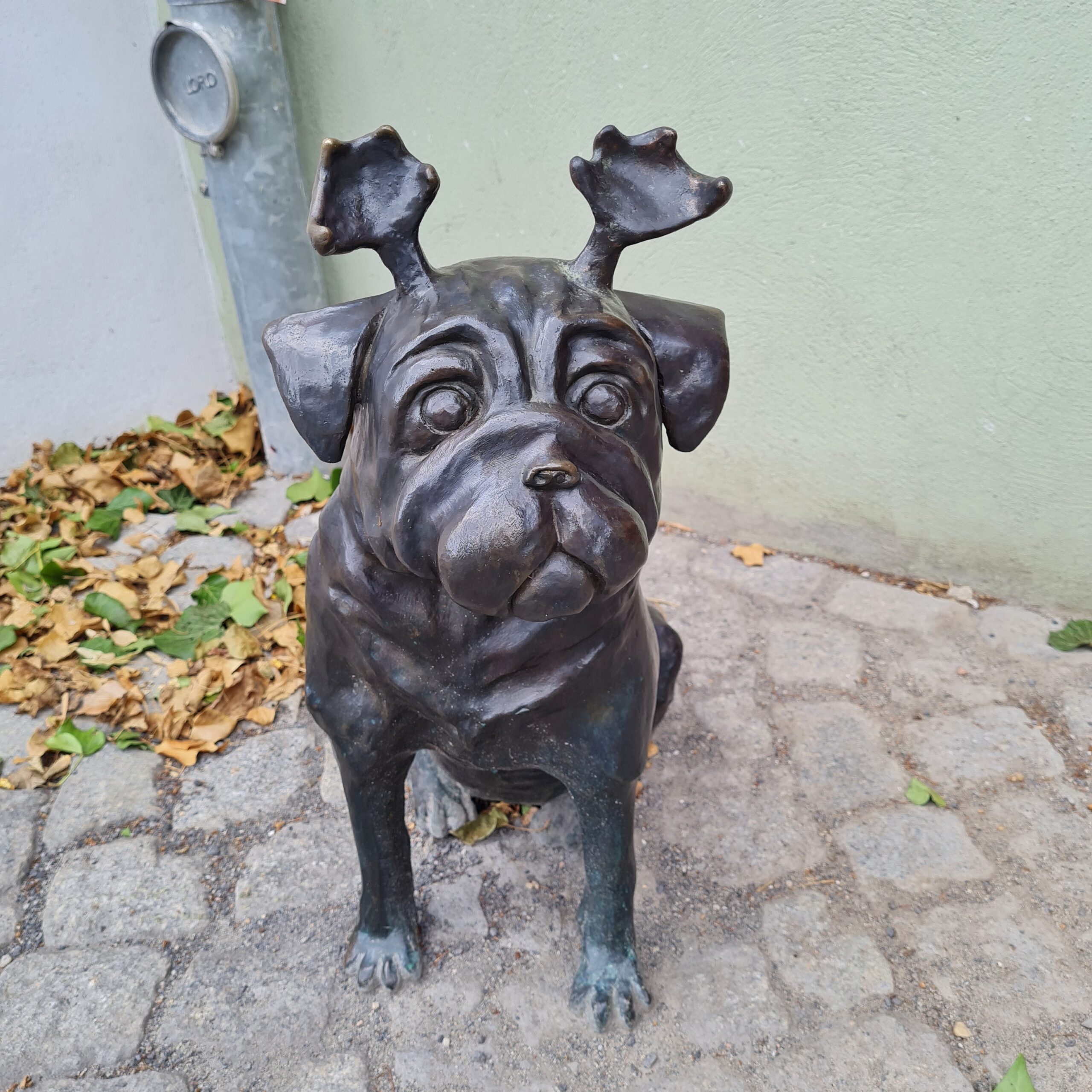 Ein sitzender Waldmops aus Metall. Er hat das Gesicht und die Statur eines Mopses, trägt aber ein kurzes Geweih.