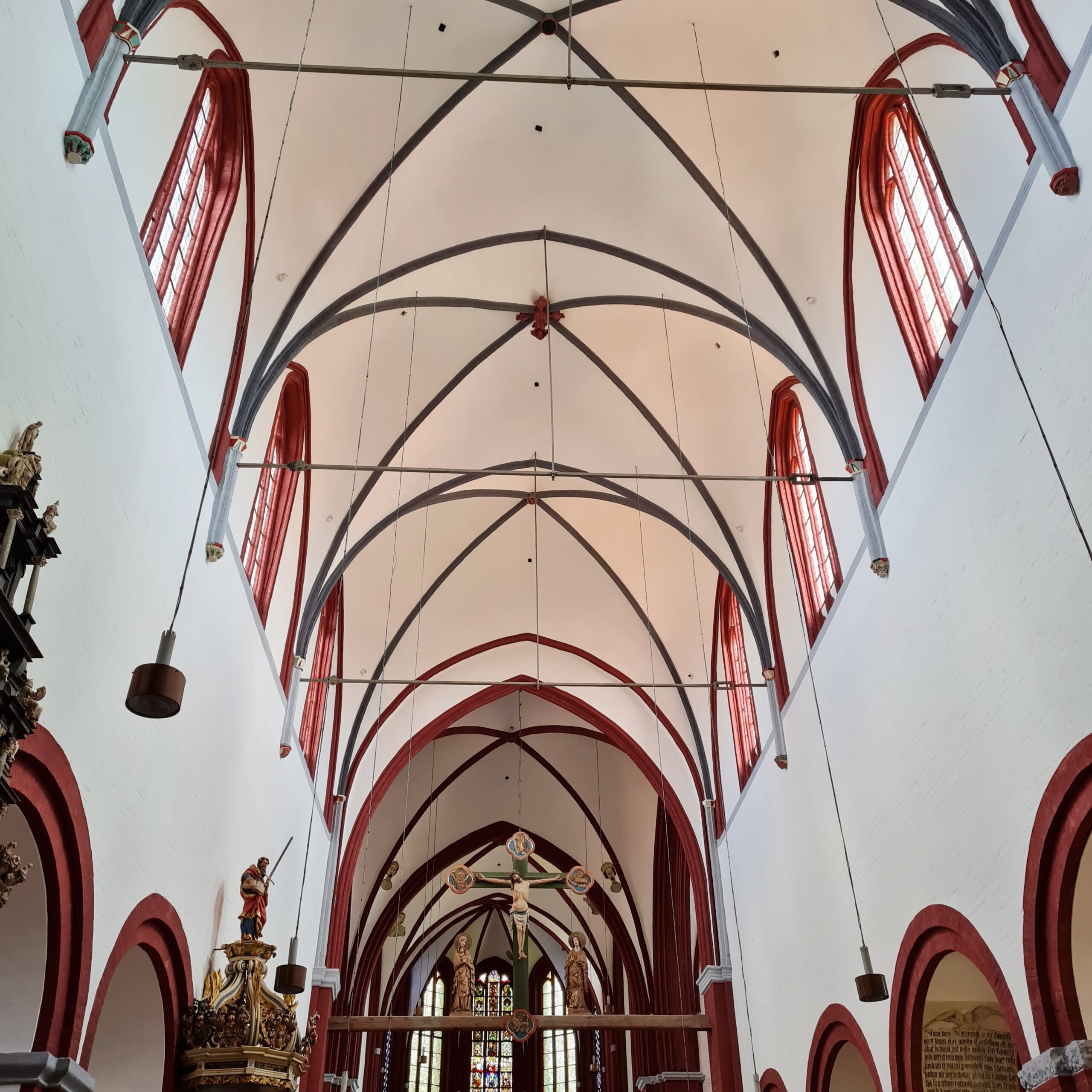Das gotische Deckengewölbe. Es ist weiß. Die Fenster haben rötliche Umrandungen und die Bögen schwarze.