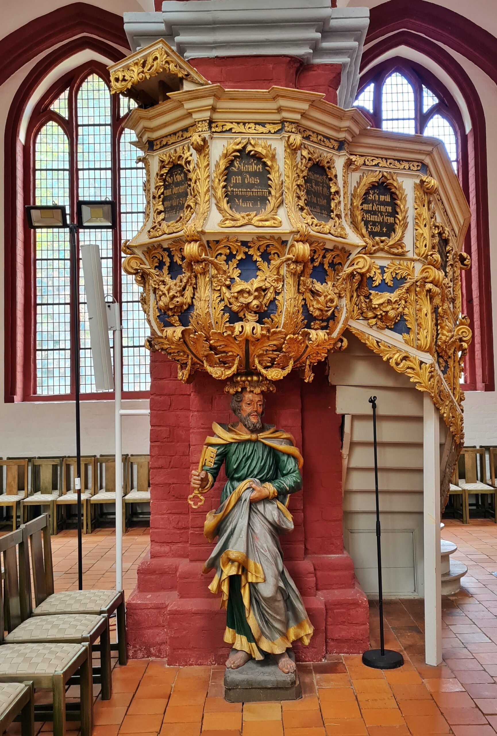 Die Kanzel, die en einem roten Kirchenpfeiler angebracht ist. Sie ist mit viel Gold verziert. Außerdem sind auf runden Platten Bibelzitate. Unter der Kanzel steht eine männliche Statue, so das es aussieht, als ob sie die Kanzel auif dem Kopf trägt. Der bärtige Mann ist barfuss. Er trägt ein weites dunkelgrünes Gewand mit goldenen Borten. Mit der linken Hand rafft er es nach oben, so dass die graue Unterseite des Gewandes sichtbar wird. In der anderen Hand hält er einen großen goldenen Schlüssel.