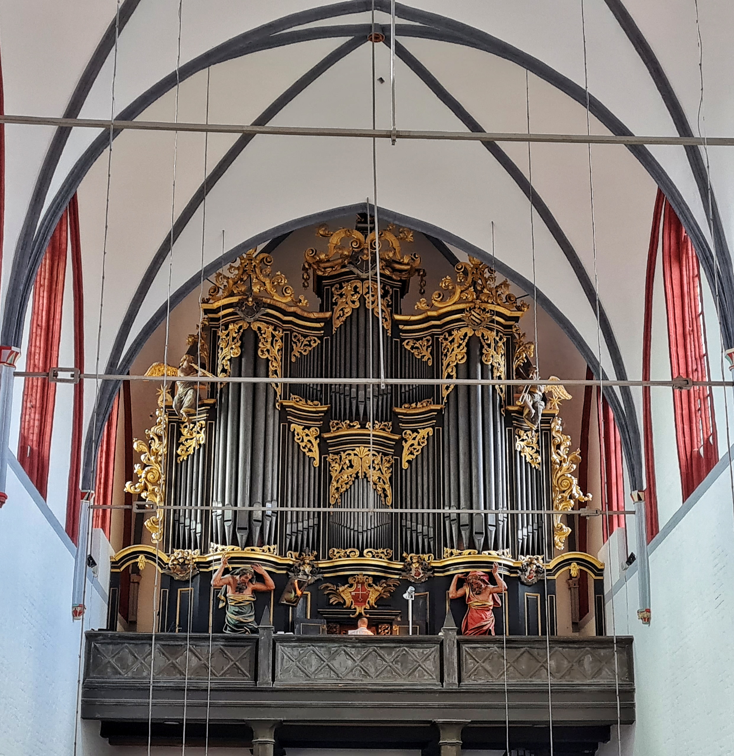 Eine große dunkle Orgel mit goldenen Verzierungen. Man sieht den Oberkörper des Organisten von hinten. Links und rechts davon stehen zwei große männliche Statuen in weiten Gewänder (links grün, rechts rot). Sie haben die Hände abgewinkelt erhoben, so das es aussieht als ob sie die Orgel tragen.