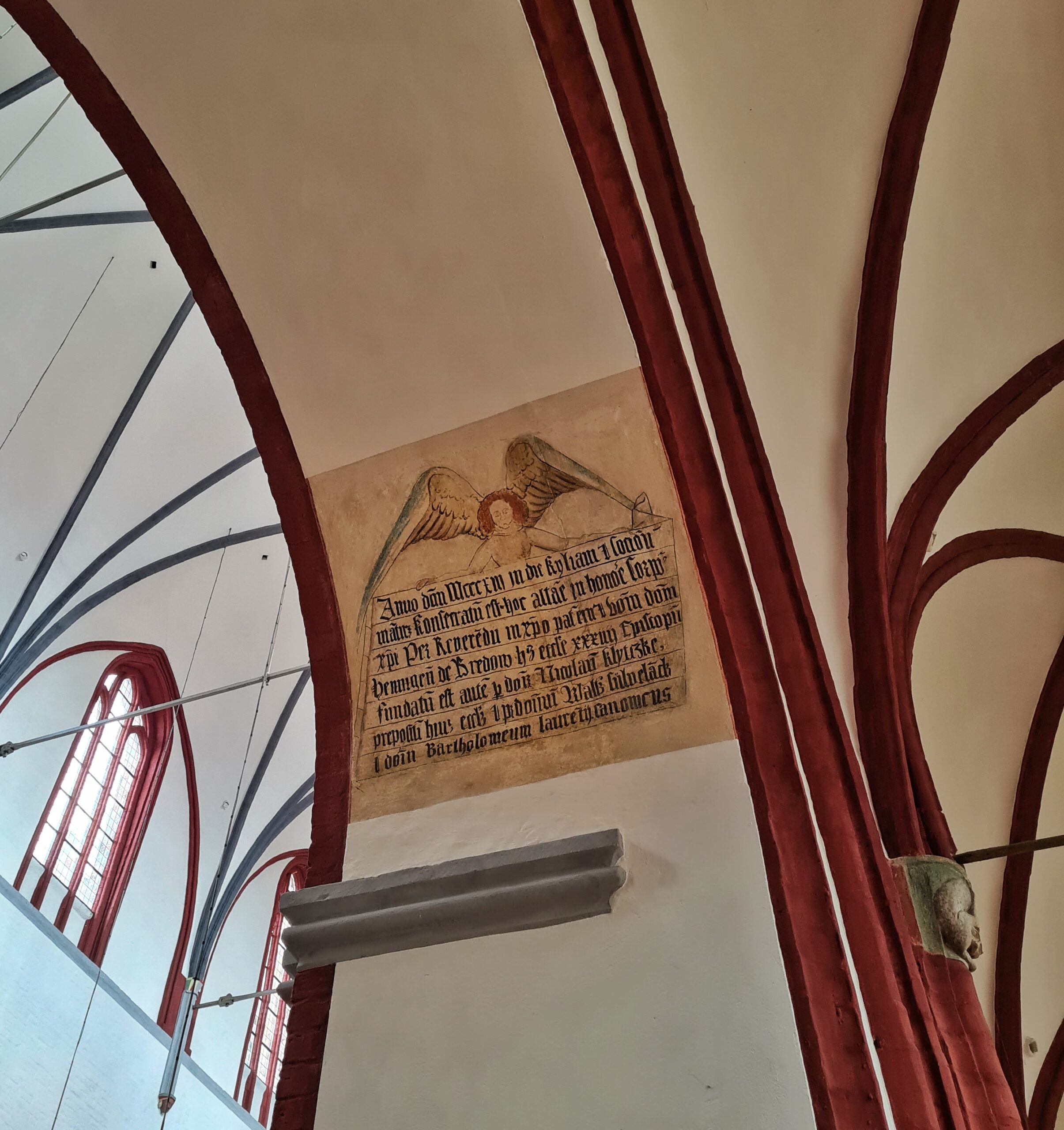 Der Bogen eines Pfeilers (weiß mit rötlichen Umrandungen) Darauf ist eine Zeichnung eines Engels, der eine Art Transparent in den Händen trägt. Darauf Text in mittelalterlicher Sprache.