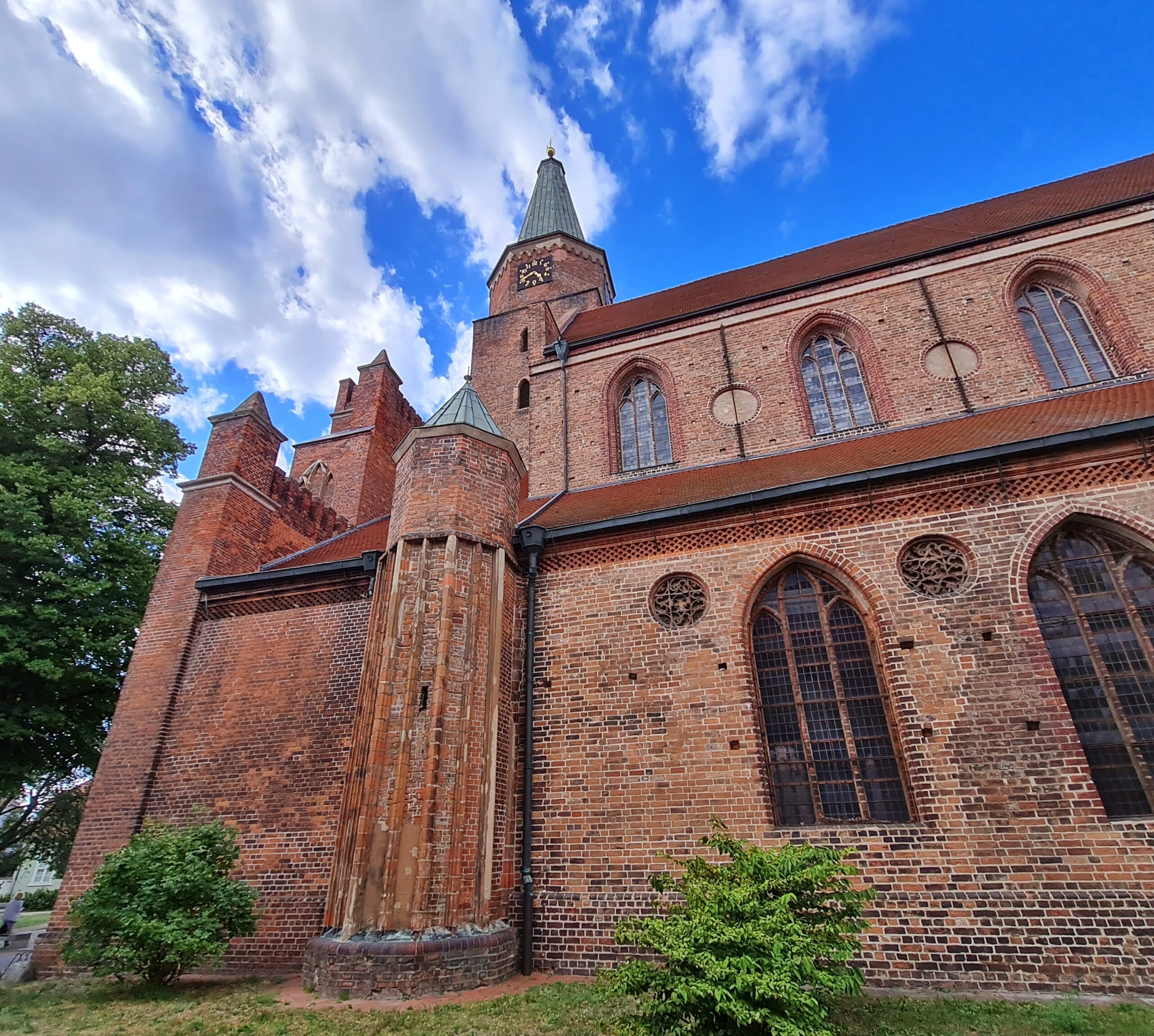 Ein Teil einer großen Backsteinkirche, seitlich schräg fotografiert. Die Kirchenfenster oben und unten sind länglich undn nach oben halbrund. Ein kleiner Turm ist außen am Gebäude angebracht. Der Kirchturm ragt darüber in den Himmel.