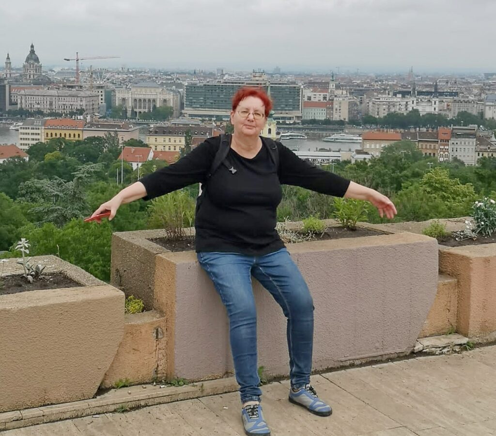 Eine Frau sitzte auf dem Rand eines hohen Blumenkübels. Im Hintergrund die Bebauung Budapests und die Donau. Die Frau hat rote Haare und eine Brille, Sie trägt Jeans und ein schwarzes T-Shirt. Sie hat die Arme leicht ausgebreitet.
