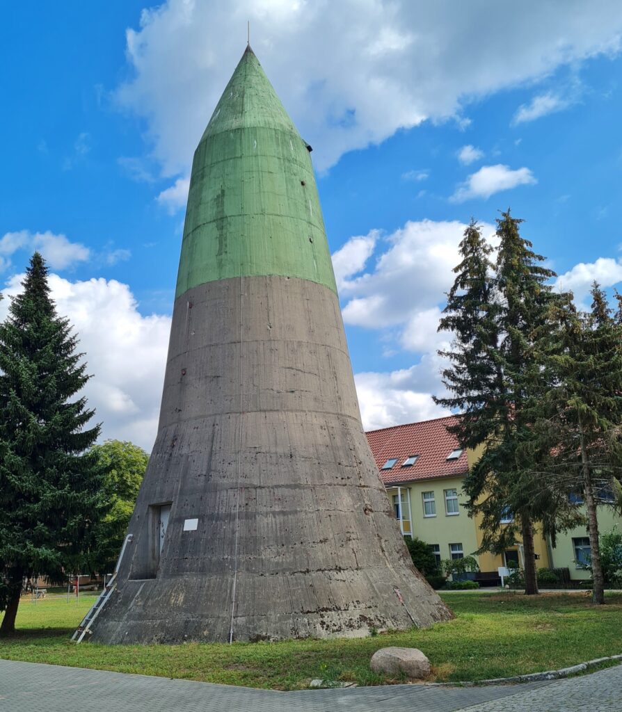 Vor einem zweigeschossigen Wohnblock steht auf einer Grünfläche mit Bäumen ein hoher, nach oben spitz zulaufender Turm. Er ist im unteren Teil grau und im oberen hellgrün gestrichen. Unten führt eine kleine Metallleiter zu einer geschlossenen Metalltür. Ansonsgten hat der Turm keine Öffnungen.
