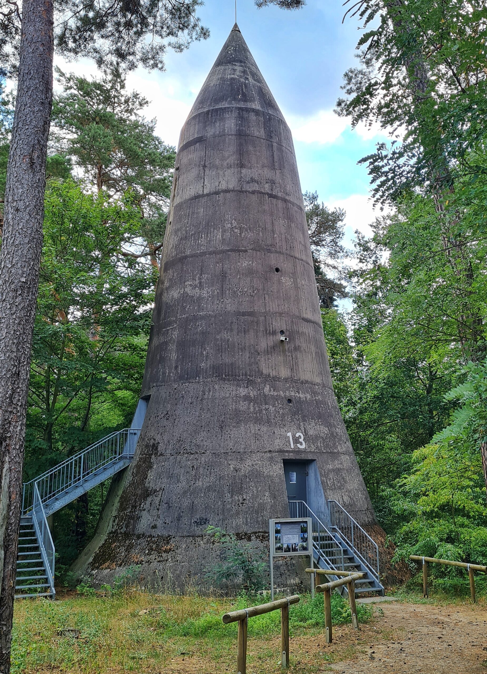 Noch einer der oben spitz zulaufenden Türme, diesmal ganz in grauem Beton. Unten rechts geht eine kleine Metalltreppe zu einer verschlossenen Metalltür hinauf. Daneben steht eine Schautafel. Links geht eine längere Metalltreppe zu einem weiteren Eingang hinauf.
