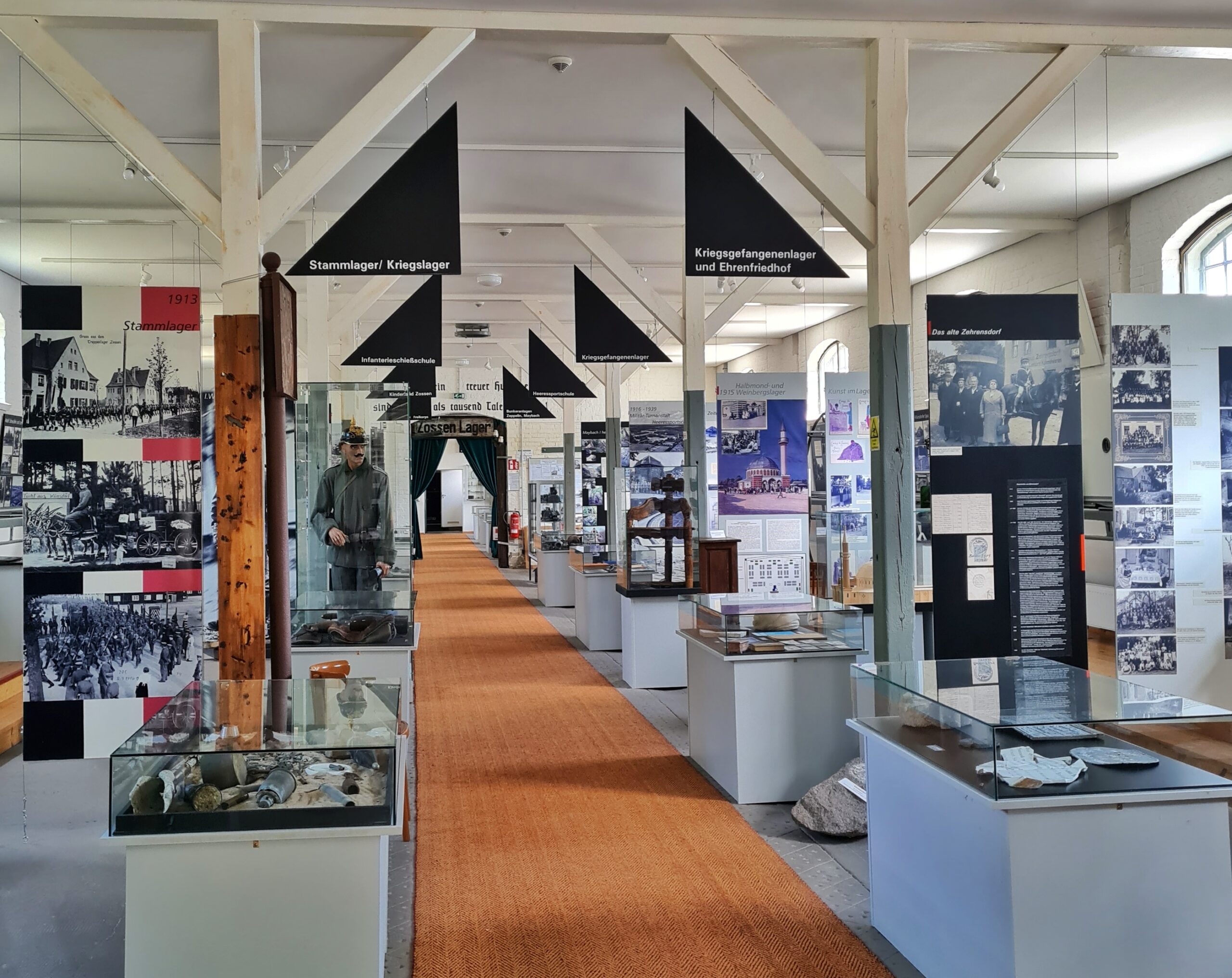 Ein Ausstellungsraum. Links und rechts vom Gang in der Mitte Vitrinen mit Ausstellungsstücken und Tafeln mit Fotos und Erklärungen.