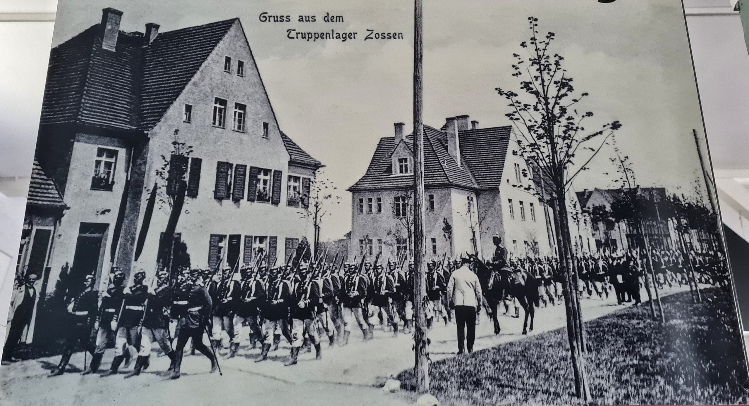 Eine alte Schwarz-Weiß-Postkarte. Vor hohen Wohnhäusern läuft eine Kolonne von Soldaten mit Gewehren auf der Schulter. Rechts von ihnen ein Soldat auf einem Pferd und ein Mann von hinten. Oben der Trxt "Gruß aus dem Truppenlager Zossen".