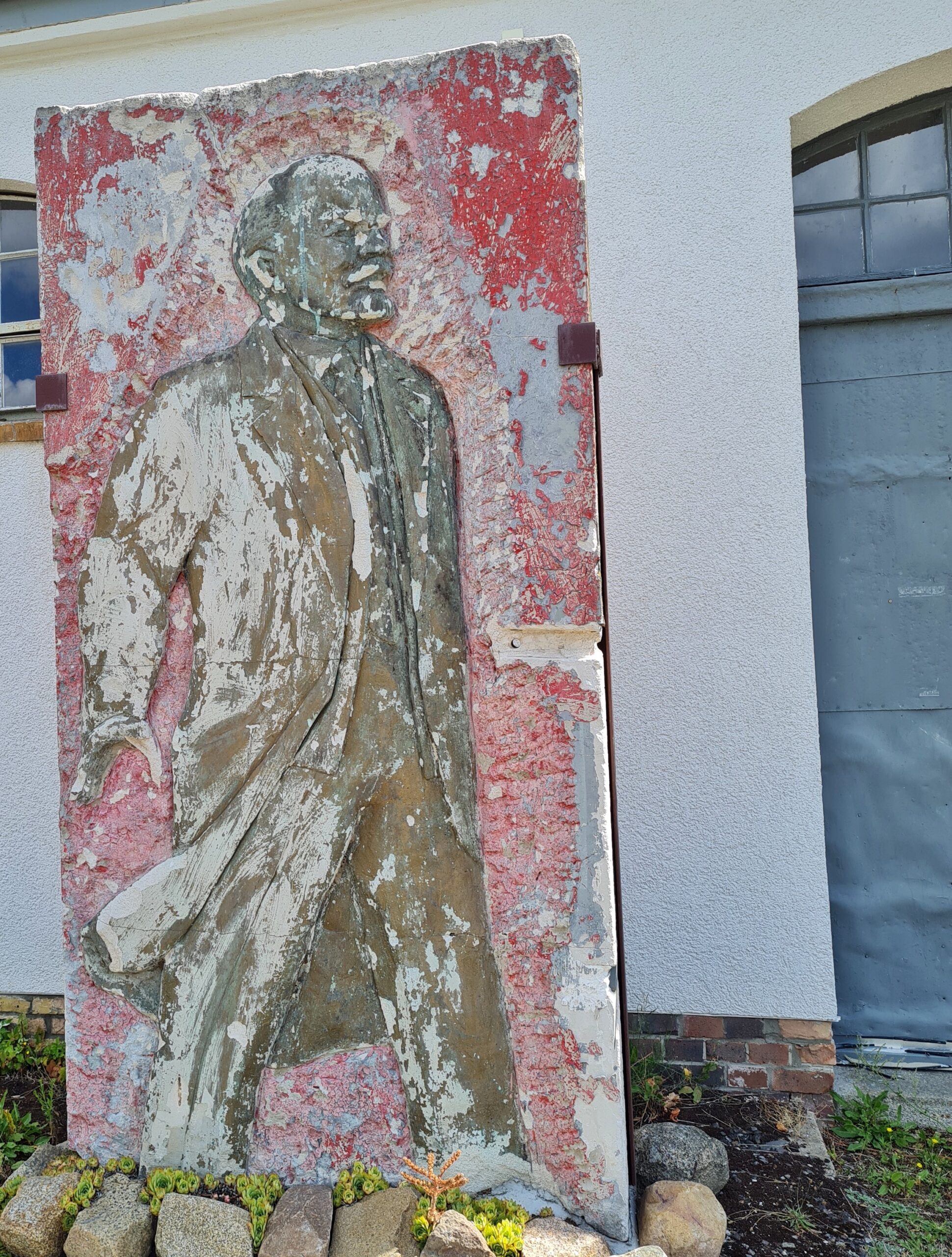 Eine rötliche hohe Platte, aufgestellt vor einem hellen Gebäude. Darauf ein Relief des gehenden Lenin.