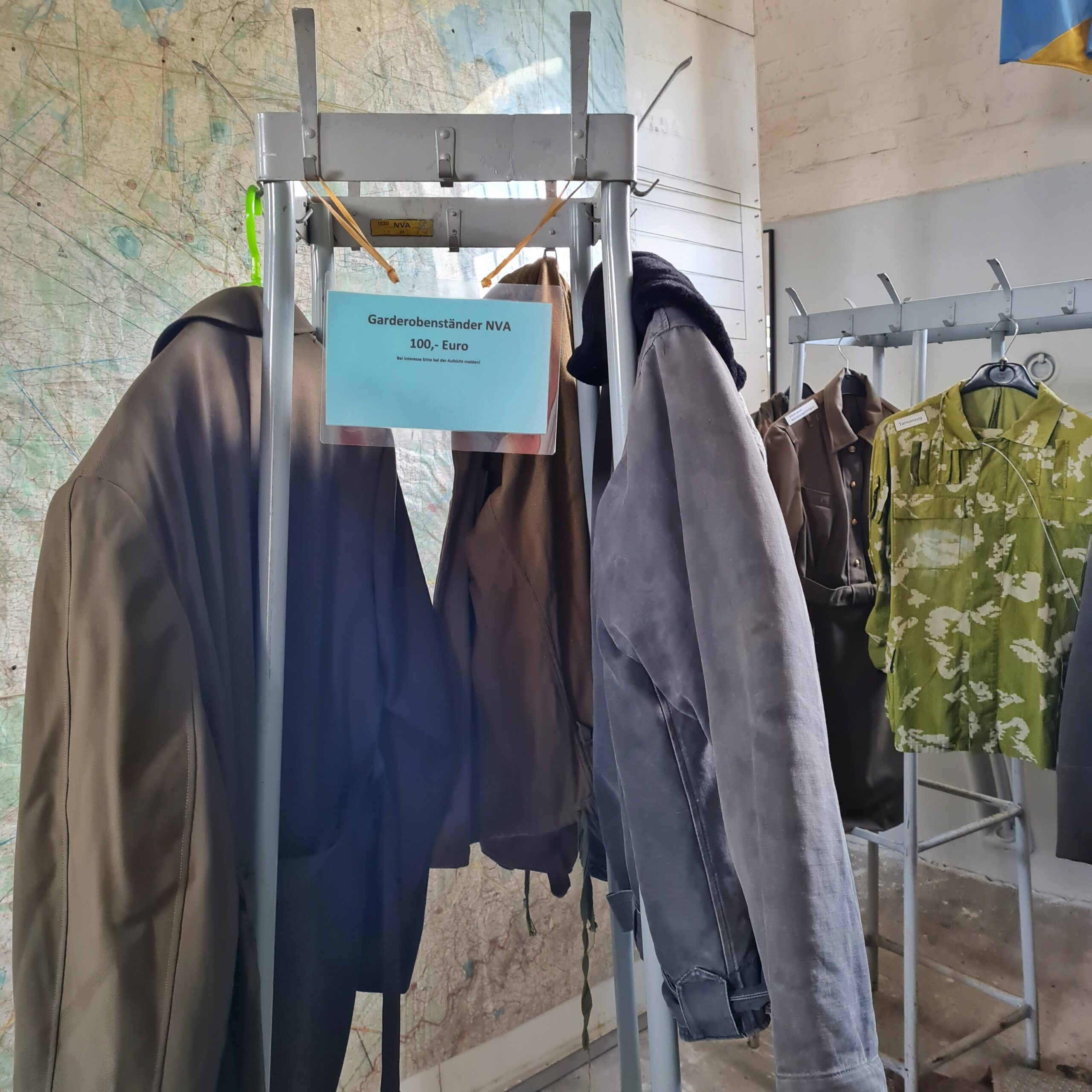 Ein viereckiger Metallständer, an dem mehrere Jacken hängen. Außerdem ein Schild: Garderobenständer NVA 100 €. Im Hintergrund ein länglicher Garderobenständer, an dem ein Mantel und ein grün-weiß-gemustertes Hemd hängt.