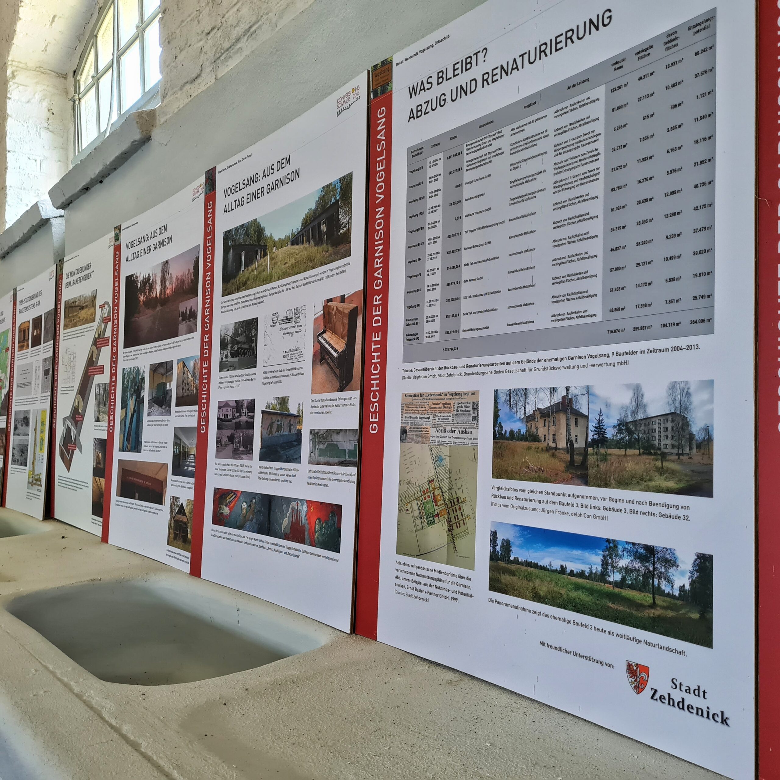 Ausstellungstafeln an einer hellen Wand, seitlich fotografiert. Darauf viele Fotos und Erklärungen. Auf den Tafeln, die am dichtesten an der Kamera sind, stehen die Überschriften "Vogelsang: Aus dem Alltag einer Garnison" und "Was bleibt? Abzug und Renaturierung".