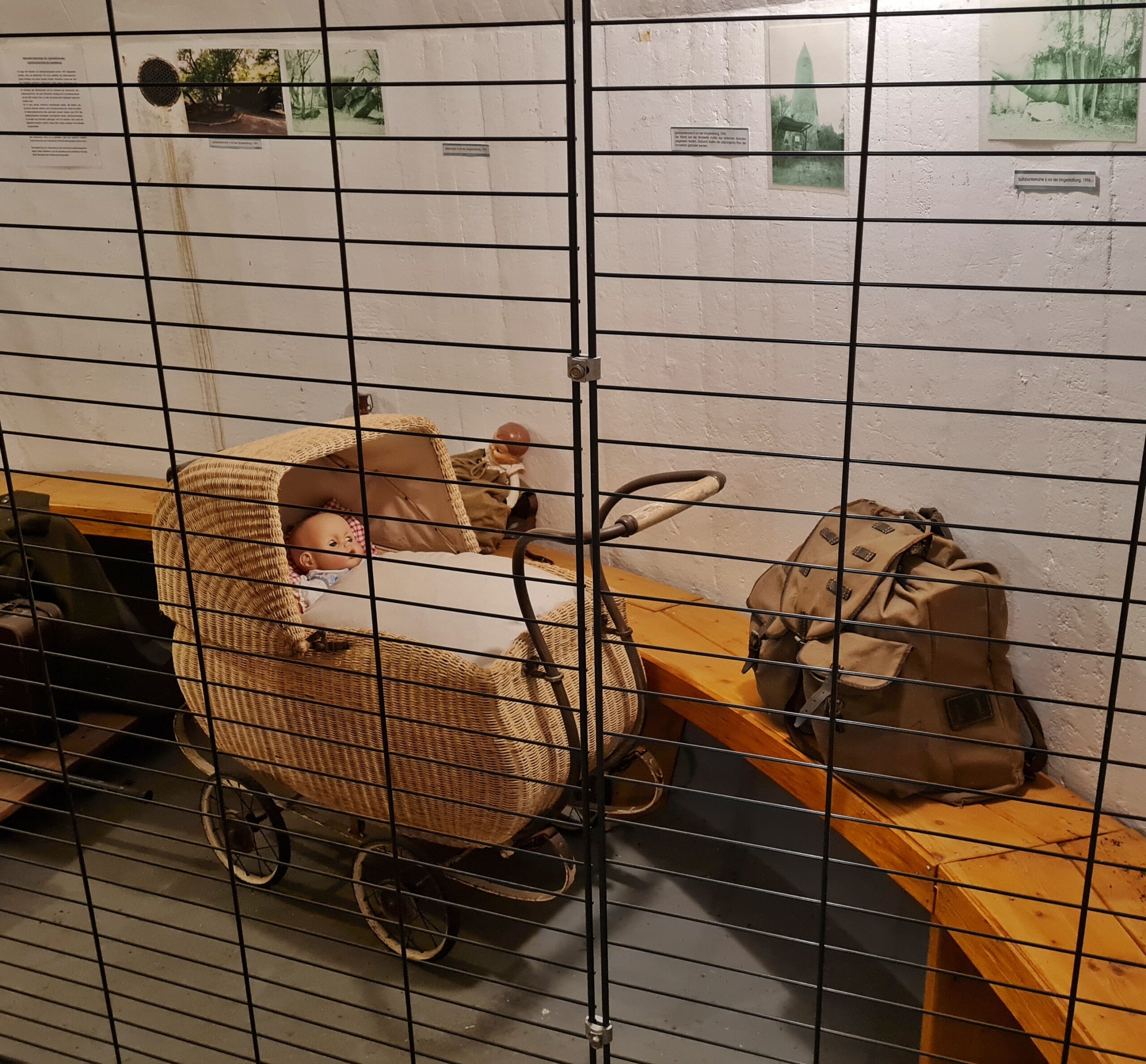 Ein Teil einer halbrunden Holzbank an einer hellen Wand. Darauf steht ein alter gepackter Rucksack. Davor steht ein alter Korbkinderwagen, in dem eine Babypuppe liegt. Davor ein Gitter.