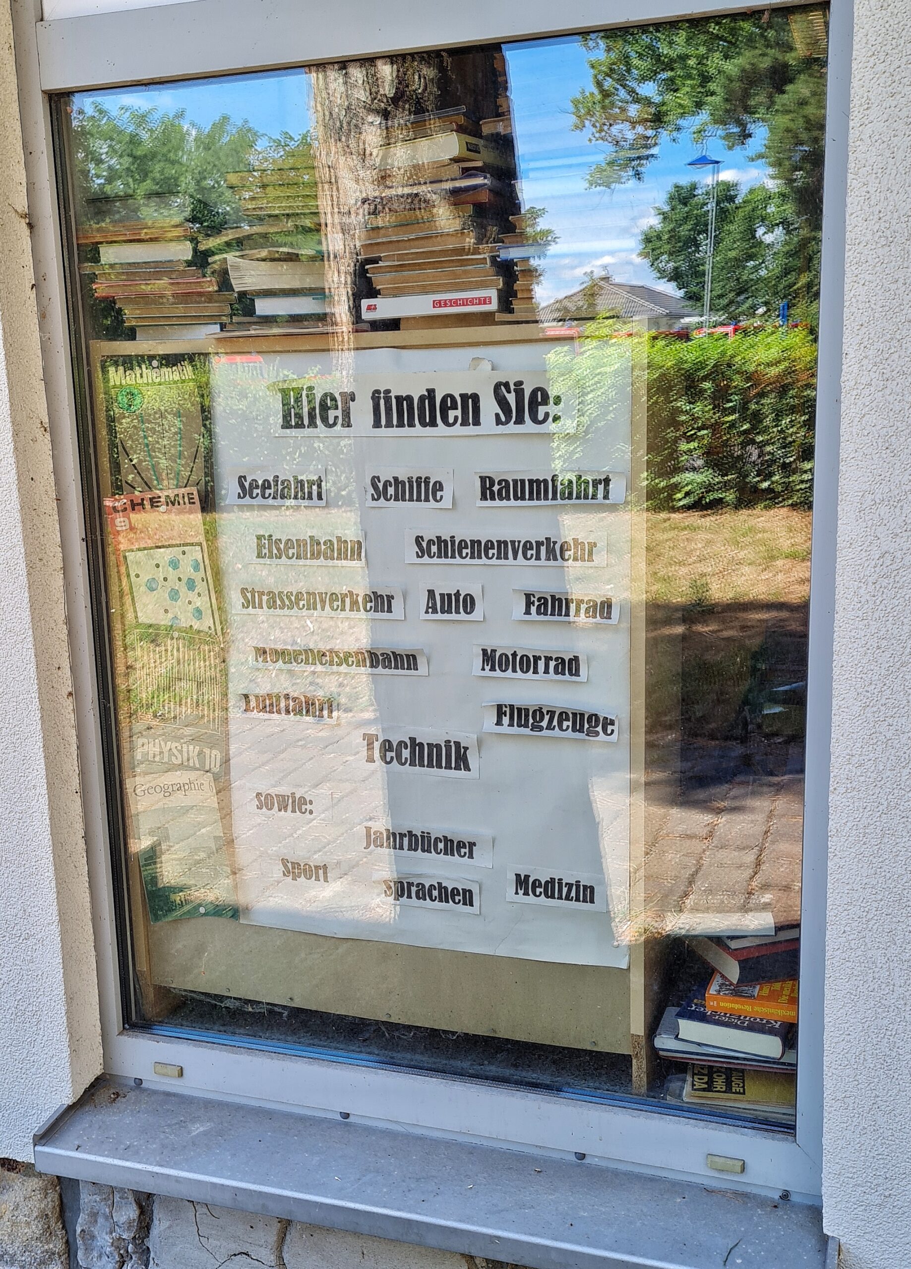 Foto eines Schaufenstes eines Antiquariats. Darin eine große Tafel: Hier finden Sie: Seefahrt Schiffe Raumfahrt Eisenbahn Schienenverkehr Strassenverkehr Auto Fahrrad Modelleisenbahn Motorrad Technik sowie Jahrbücher Sport Sprachen Medizin. Im Hintergrund aufgestapelte Bücher. In der Scheibe spiegeln sich eine gepflasterte Fläche, eine Hecke und das Dach eines Einfamilienhauses.
