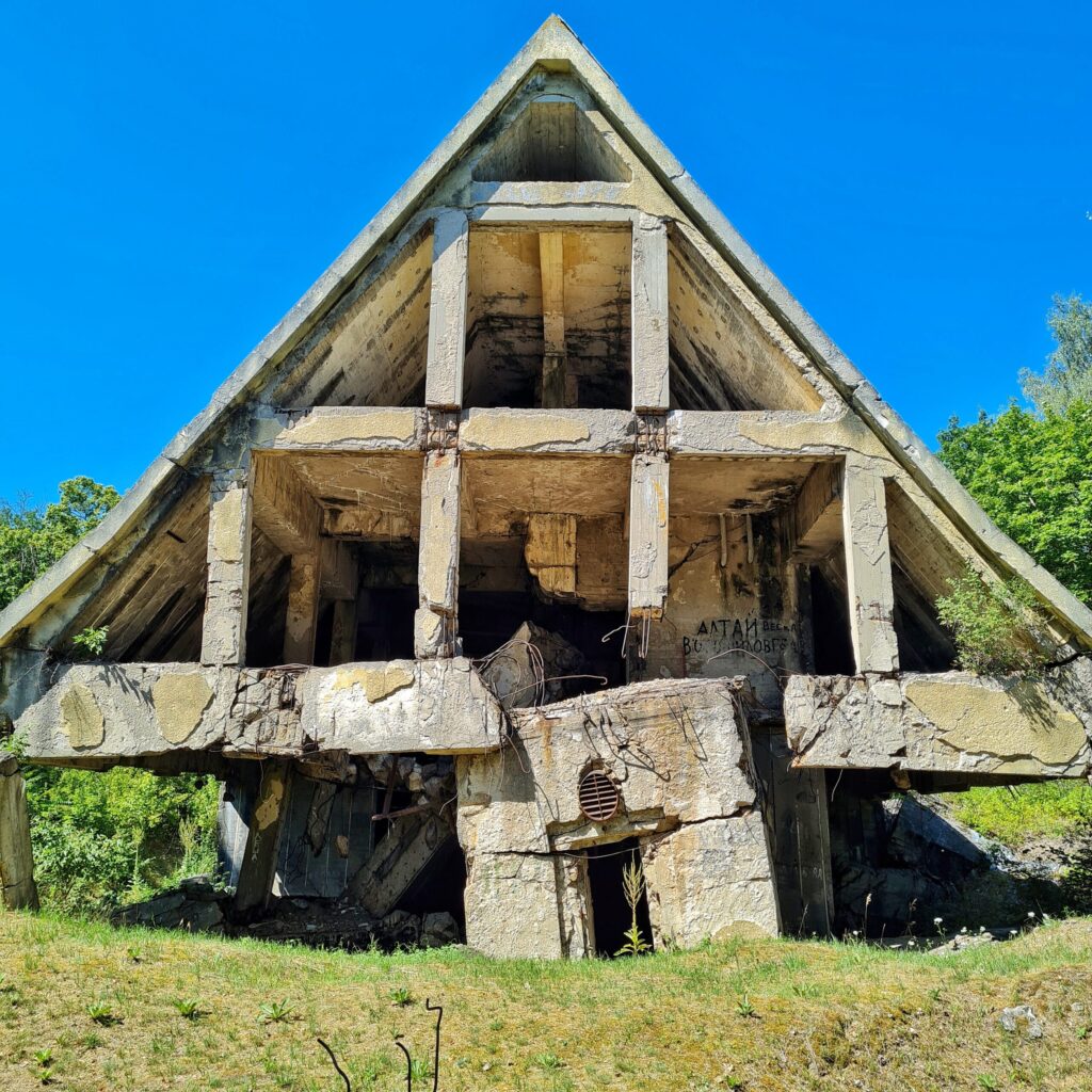 Ein zusammengestürztes Gebäude. Auf den Trümmern das stehende Spitzdach, nach vorne offen, so dass man die früheren Räume darin sehen kann. 