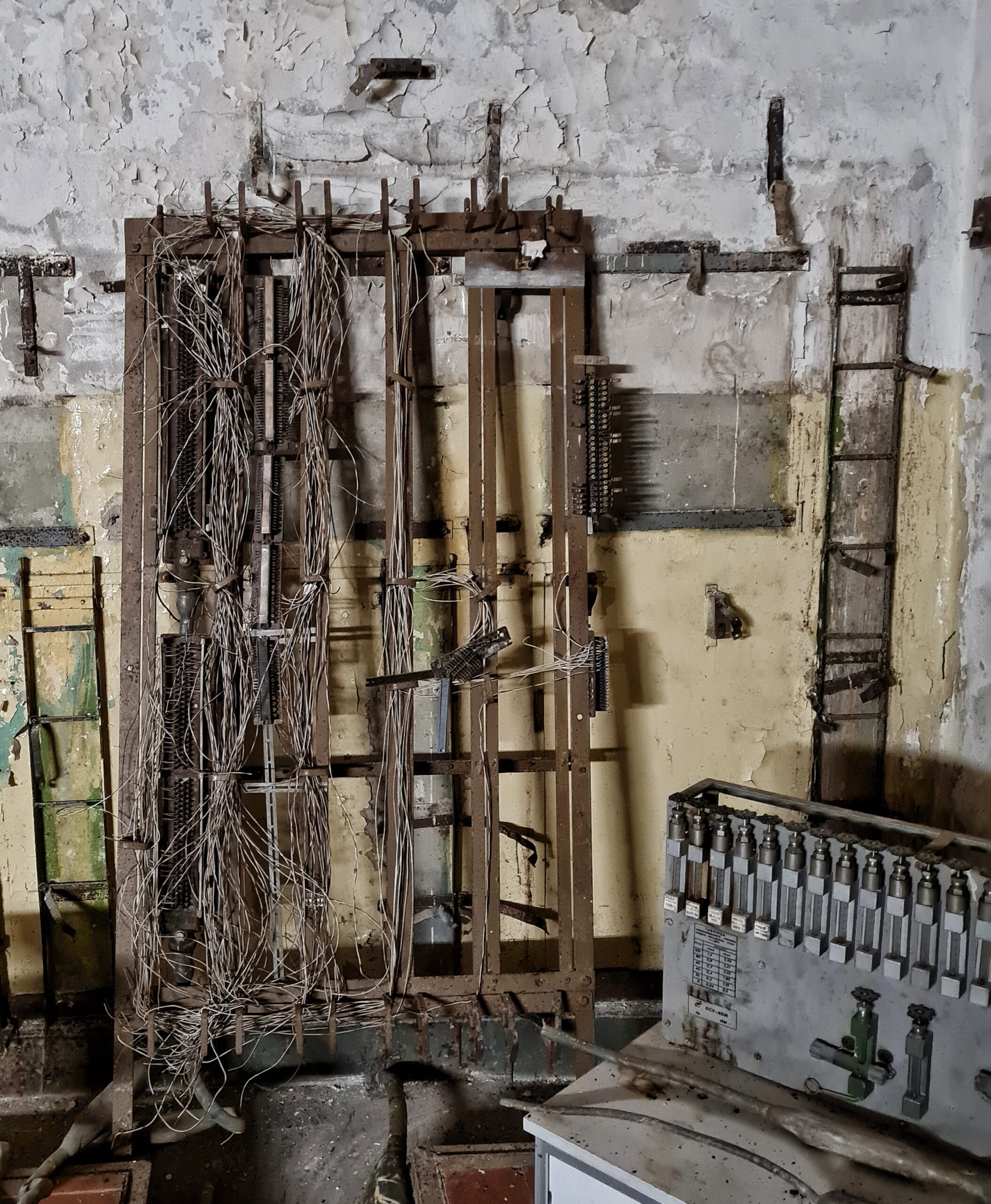 An eine Mauer gelehnt ein Metallgitter mit senkrechten Streben. Daran hängen zusammengerollt Kabel herunter. Rechts ein Gerät mit Schaltern.