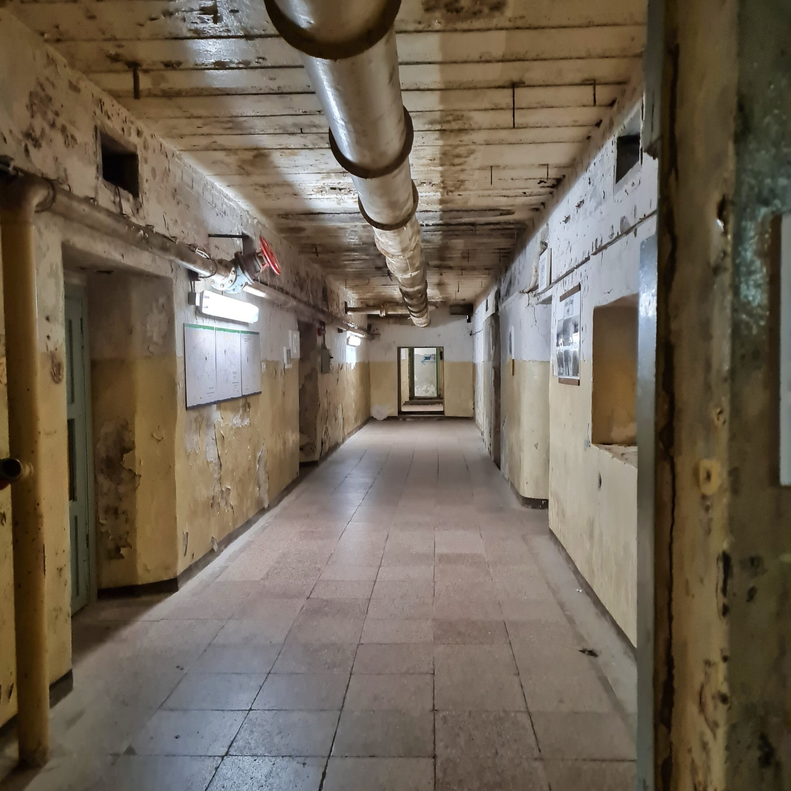 Ein langer Flur mit gepflastertem Boden. Das einzige Licht kommt von einer Lampe an der linken Wand. An der Decke verläuft in der Mitte ein großes Rohr. Links und rechts Türffnungen.