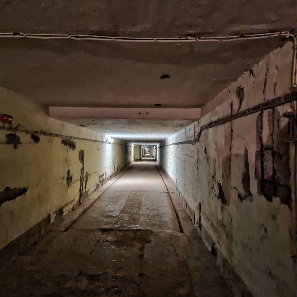 Ein sehr langer viereckiger Tunnel. Licht kommt nur von in großen Abständen an einer Wand angebrachten Lampen.
