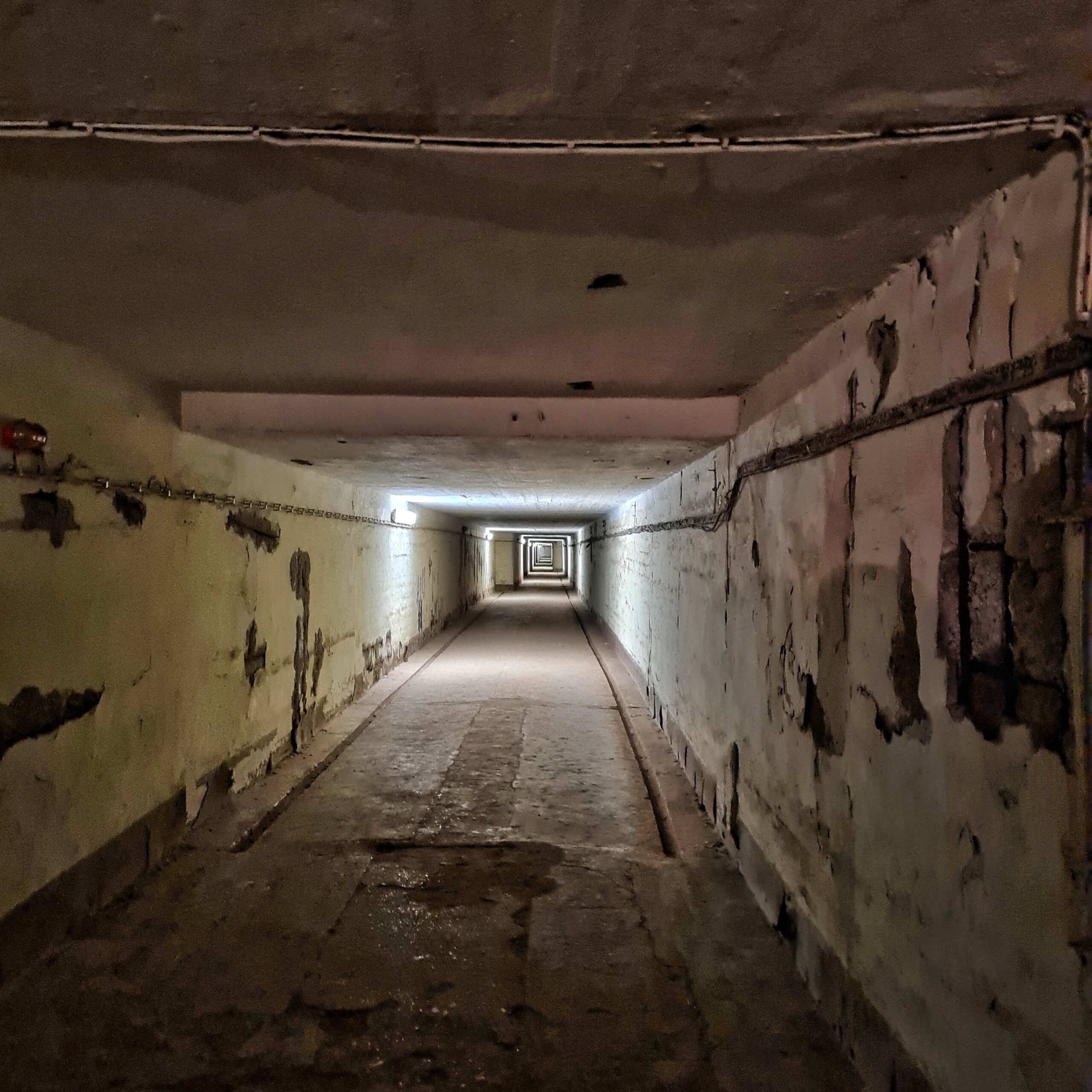 Ein sehr langer viereckiger Tunnel. Licht kommt nur von in großen Abständen an einer Wand angebrachten Lampen.