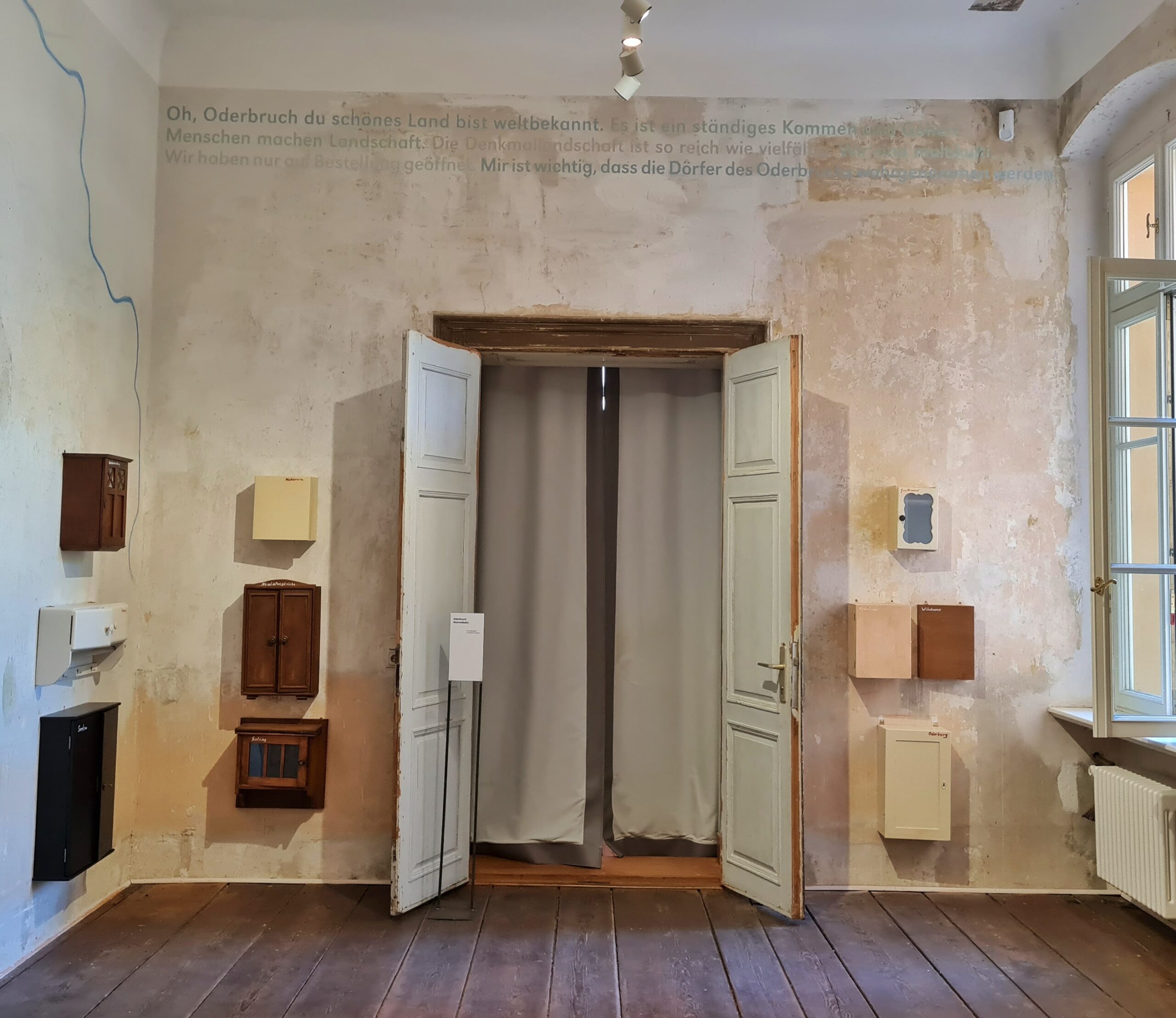 Ein Teil eines hohen Raumes mit weißlichen Wänden und einem Holzfußboden. An der hinteren Wand eine geöffnete hohe Tür, die mit einem Vorhang verdeckt ist. Links und rechts davon und ebenfalls an der linken Wand hängen kleine geschlossene Holzchränkchen, die alle völlig unterschiedlich sind. Rechts ein halb geöffnetes hohes Fenster.