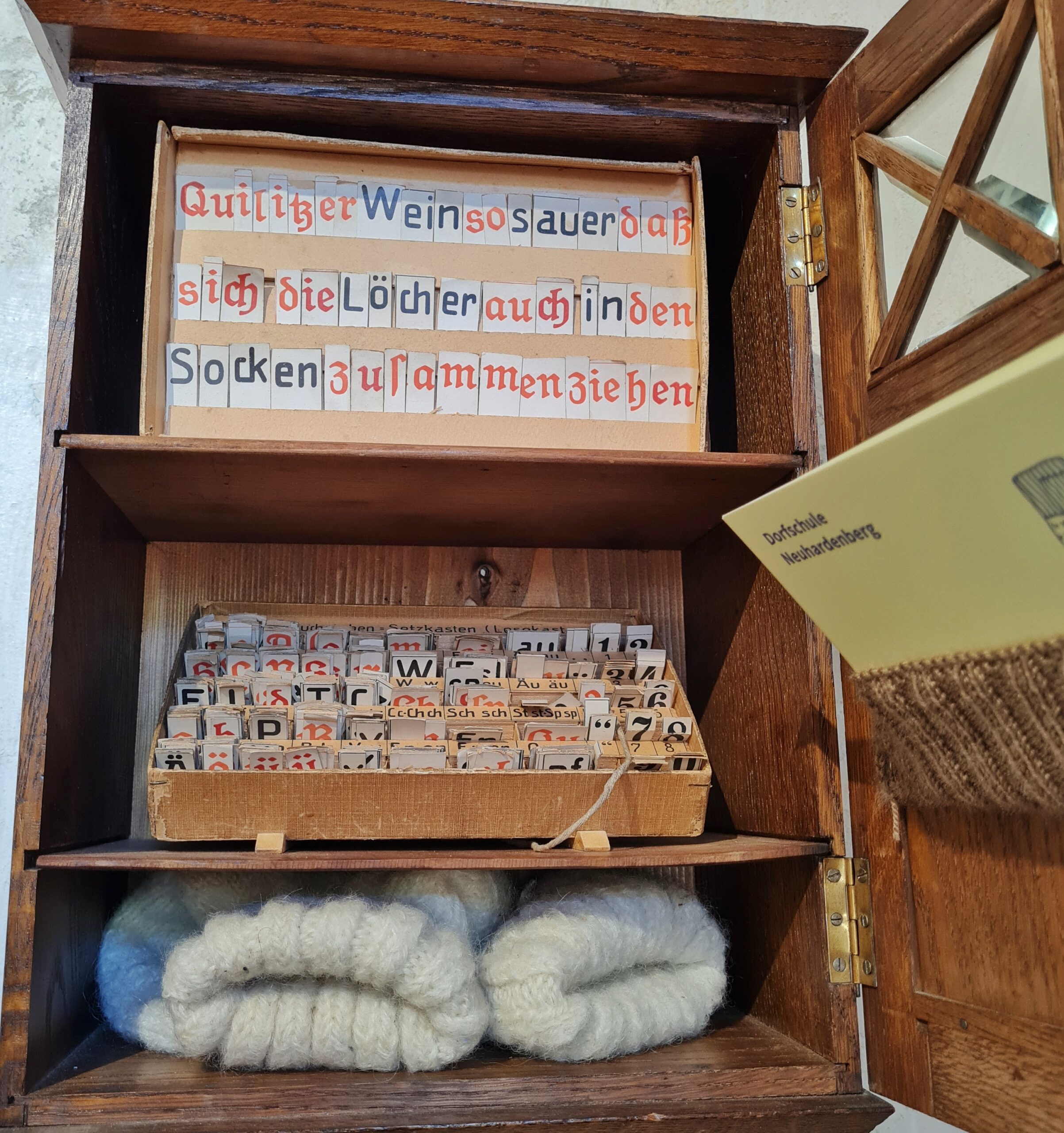 Ein geöffnetes Schränkchen mit drei Fächern, Im unteren liegen zwei weiße Wollsocken. Im mittleren steht eine Schachtel mit vielen einzelnen Buchstaben aus Papier. Im oberen sind auf einen Pappkarton Buchstaben aufgeklebt und bilden den Satz "Quliitzer Wein so sauer daß sich die Löcher auch in den Socken zusammenziehen". Rechts ein Teil des Türchens, in dem ein gestricktes Täschchen hängt. Darin steckt eine Karte mit der Aufschrift "Dorfschule Neuhardenberg".