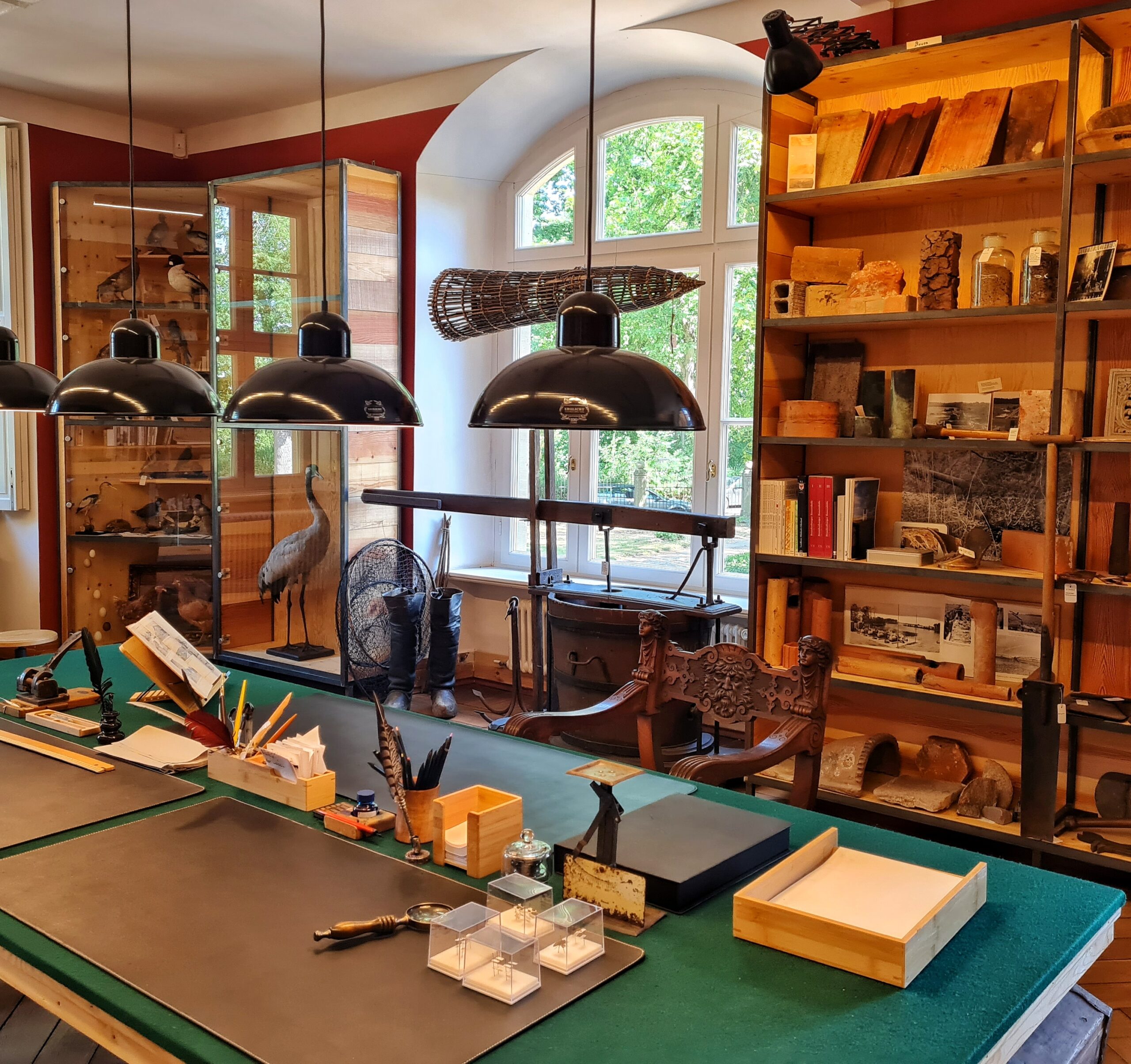 Ein Raum mit einem großen Arbeitstisch in der Mitte. Drauf Holzkästen mkit Papier und Notizzetteln, Federn in einem Holzbecher, daneben ein Tintenfass, Stifte, eine Lupe, kleine Objekte in Plexiglasbehältern. Darüber hängen drei Hängeleuchten. Hinter dem Tisch ein Holzstuhl mit Schnitzarbeiten. an der Wand neben einem großen Fenster ein Holzregal mit Büchern, Gesteins- Holz- und Bodenproben. Vor dem Fenster hohe Stiefel, ein Fischernetz, ein Anker und ein großer Holzeimer. Links davon ausgestopfte Vögel hinter Glas.