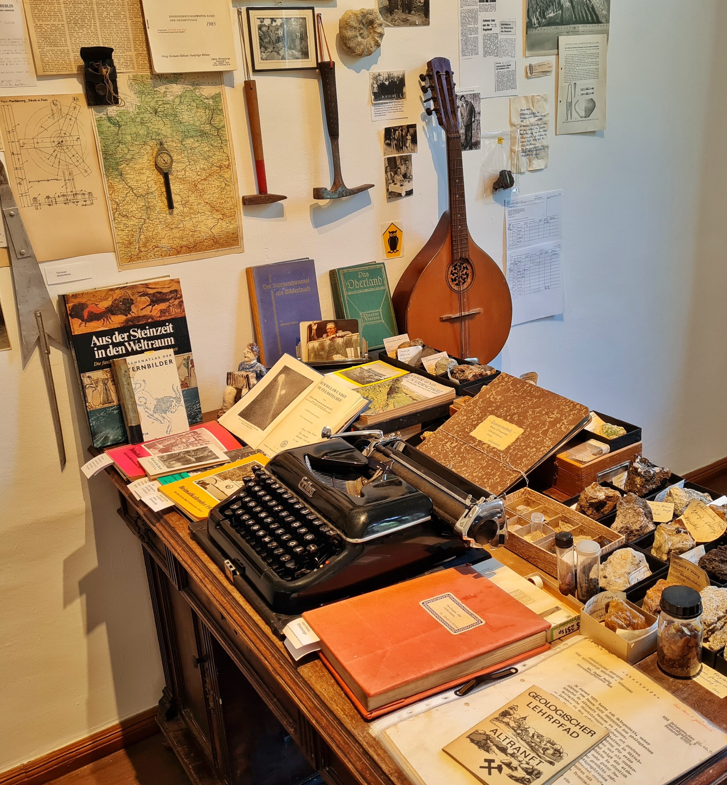 Ein Schreibtisch von der Seite fotografiert. Er ist voller Dinge: eine alte Schreibmaschine, verschiedene alte Bücher, Kästchen mit Gesteinsproben. Eine kleine Broschüre auf der rechten Seite des Schreibtisches trägt den Titel "Geologischer Lehrpfad Altranft". An der Wand eine Karte, Skizzen, Zeitungsausschnitte, Fotos, zwei Hammer und ein Zupfinstrument.