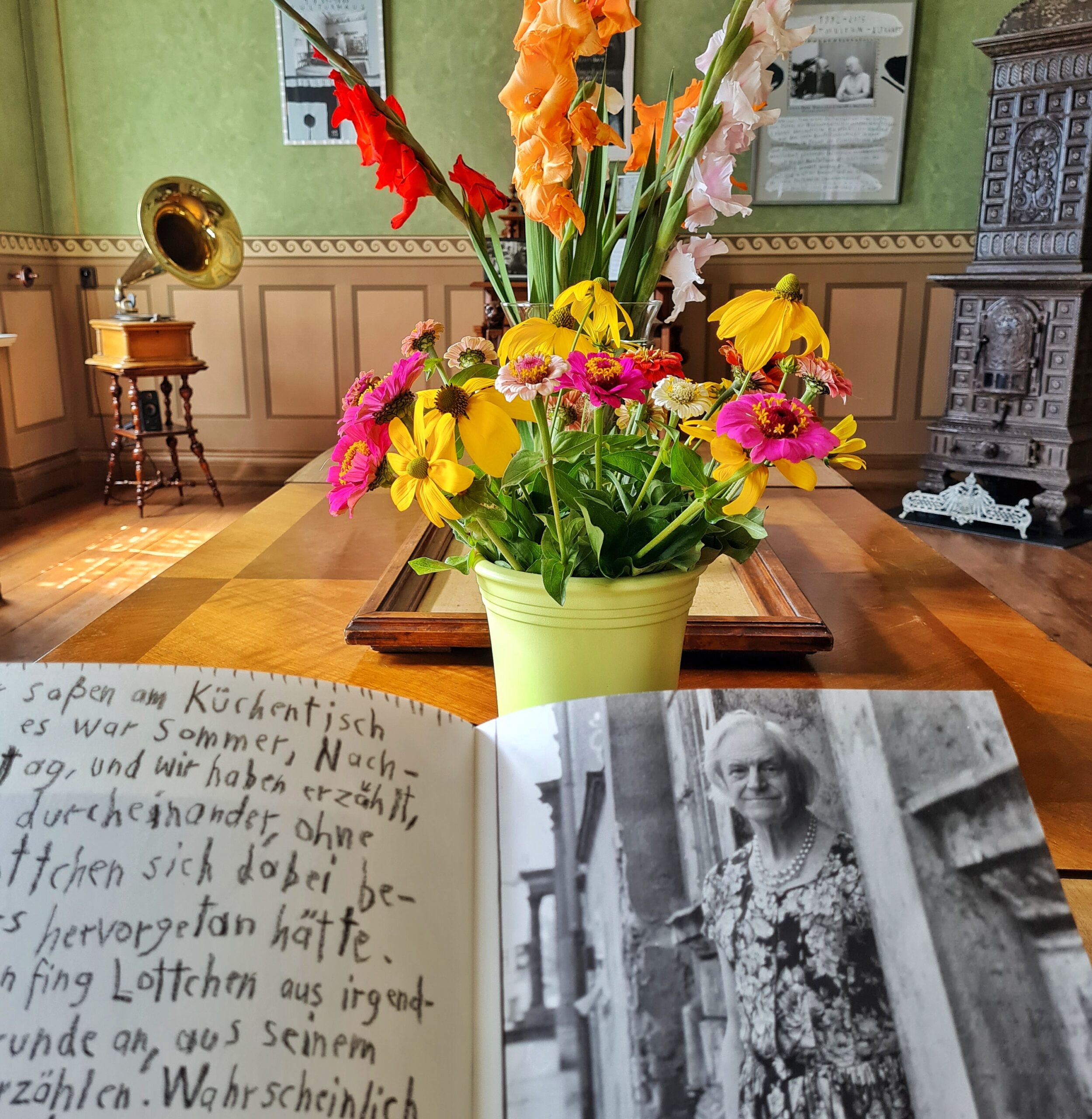 Der Tisch von der Seite fotografiert. Im Vordergrund ein geöffnetes Buch, das auf der rechten Seite in Schwarz-Weiß-Foto von Charlotte von Mahlsdorf in einem geblümten Kleid zeigt und auf der linken Seite etwas handschriftlich geschriebenes. Dahinter die Vasen mit Blumen und das Tablett. Im Hintergrund Fotos an der Wand, ein Ofen und ein Grammophon.