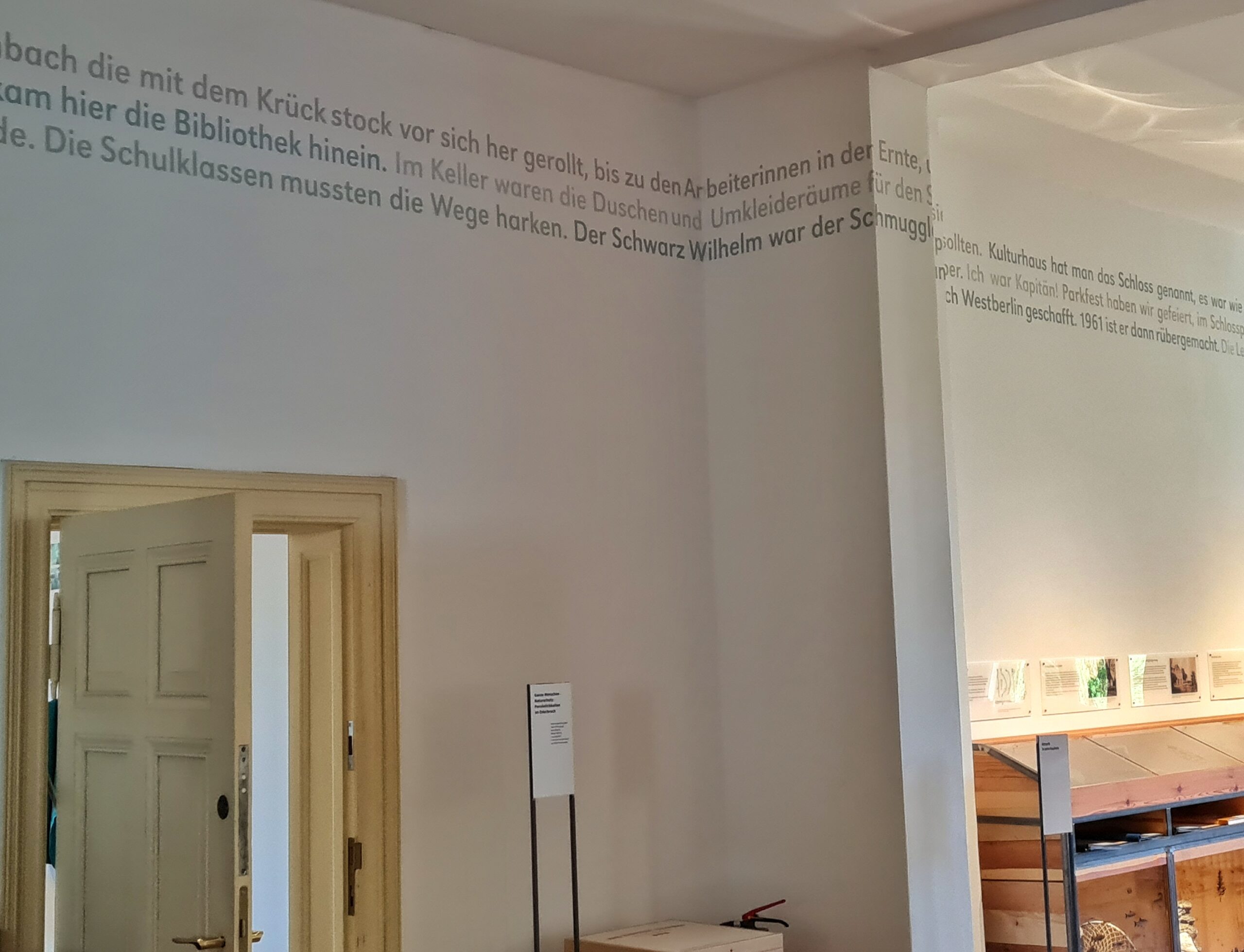 Eine hohe Zimmerwand. Unten eine geöffnete helle Holztür, eine Vitrine und zwei Informatuonstafeln. Im oberen Bereich ist durchgehend Text auf der Wand. Auf dem Foto erkennbar ist: "die mit dem Krückstock vor sich her gerollt, bis zu den Arbeiterinnen in der Ernte" und "hier die Bibliothek hinein. Im Keller waren die Duschen und Umkleideräume für den" und "Schulklassen mussten die Wege harken. Der Schwarz Wilhelm war der Schmuggler".