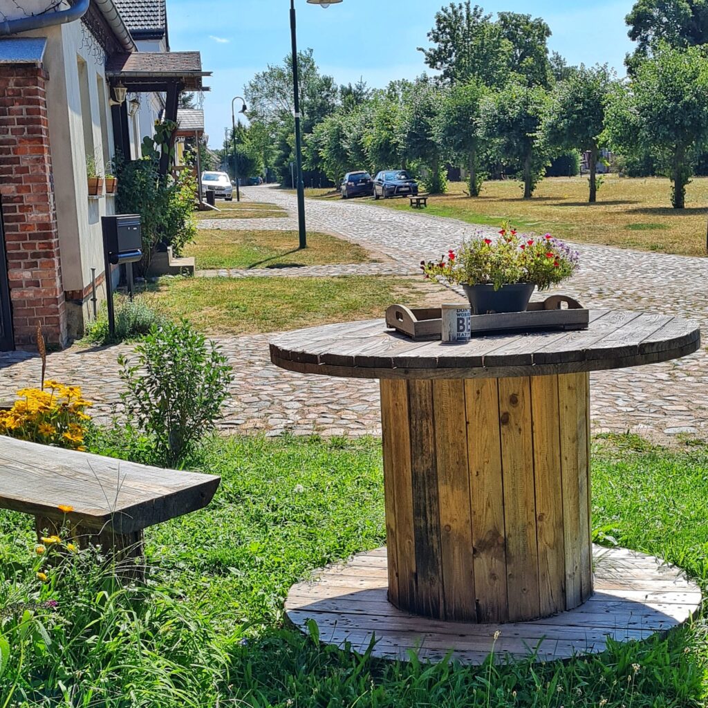 Eine gepflasterte Dorfstraße. Links sind Häuser mit Grünflächen davor, rechts Bäume in einer Reihe entlang der Straße auf einer Grünfläche. Im Vordergrund ein Holztisch aus einer Kabeltrommel. Darauf steht ein altes Holztablett mit einer Pflanze und ein Belchbehälter. Daneben ist ein Teil einer Bank zu sehen.
