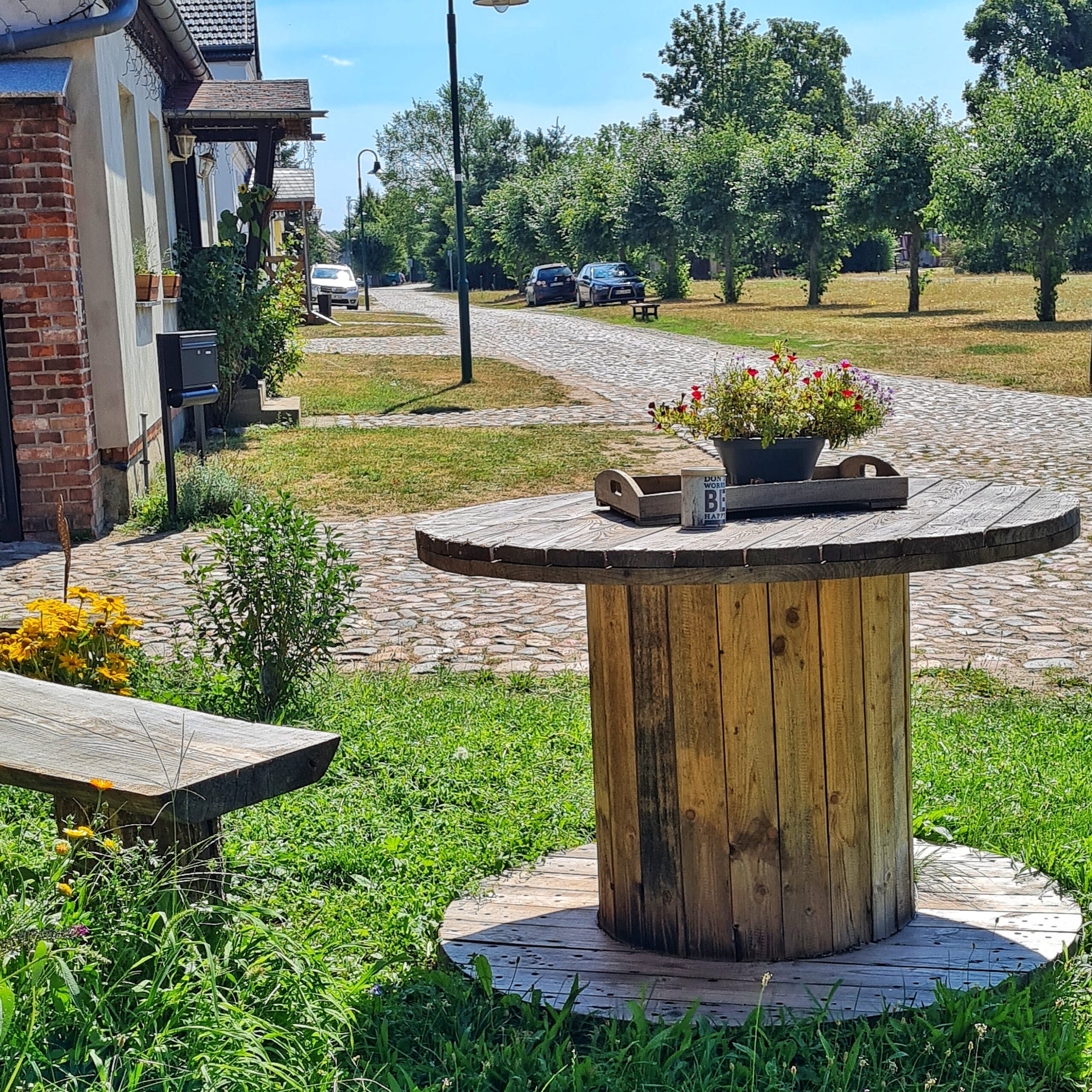 Eine gepflasterte Dorfstraße. Links sind Häuser mit Grünflächen davor, rechts Bäume in einer Reihe entlang der Straße auf einer Grünfläche. Im Vordergrund ein Holztisch aus einer Kabeltrommel. Darauf steht ein altes Holztablett mit einer Pflanze und ein Belchbehälter. Daneben ist ein Teil einer Bank zu sehen.