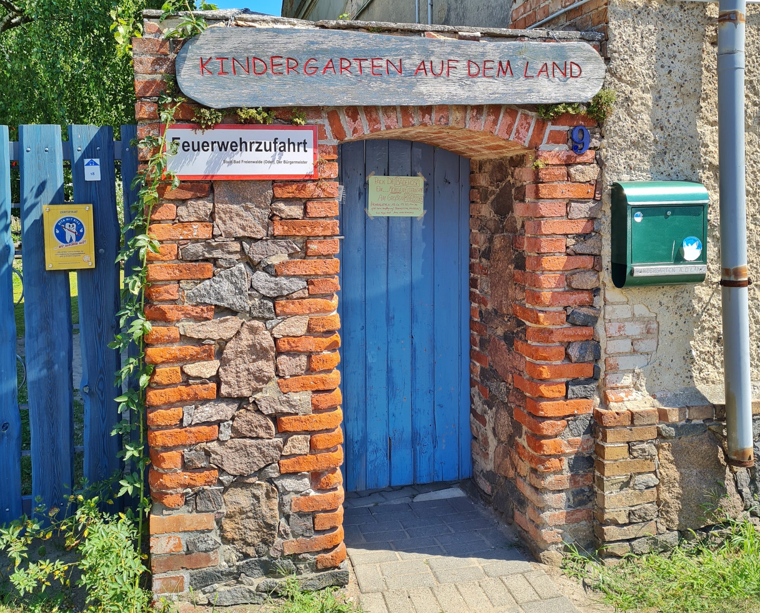 Eine Mauer mit einem blauen Holztor darin. Über dem Tor ein Holzschild: "Kindergarten auf dem Land". Rechts vom Tor ein Schild: "Feuerwehrzufahrt"